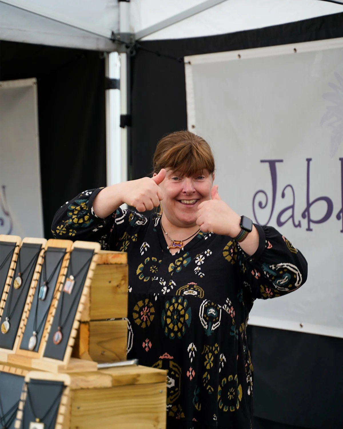 A woman smiling and giving a thumbs-up while standing behind a table with jewelry at an outdoor market or craft fair.