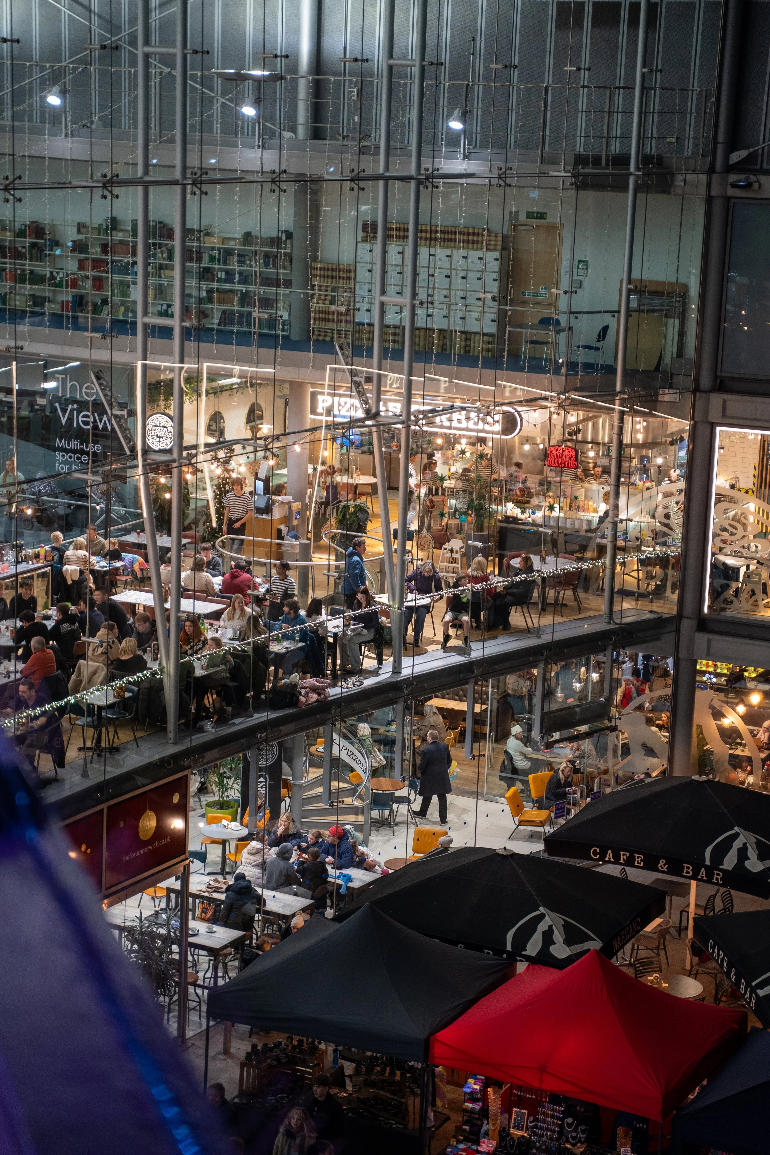 Night view of a busy multi-level indoor market or shopping mall with various stores, cafes, and seating areas visible through large glass windows, illuminated by interior lighting.