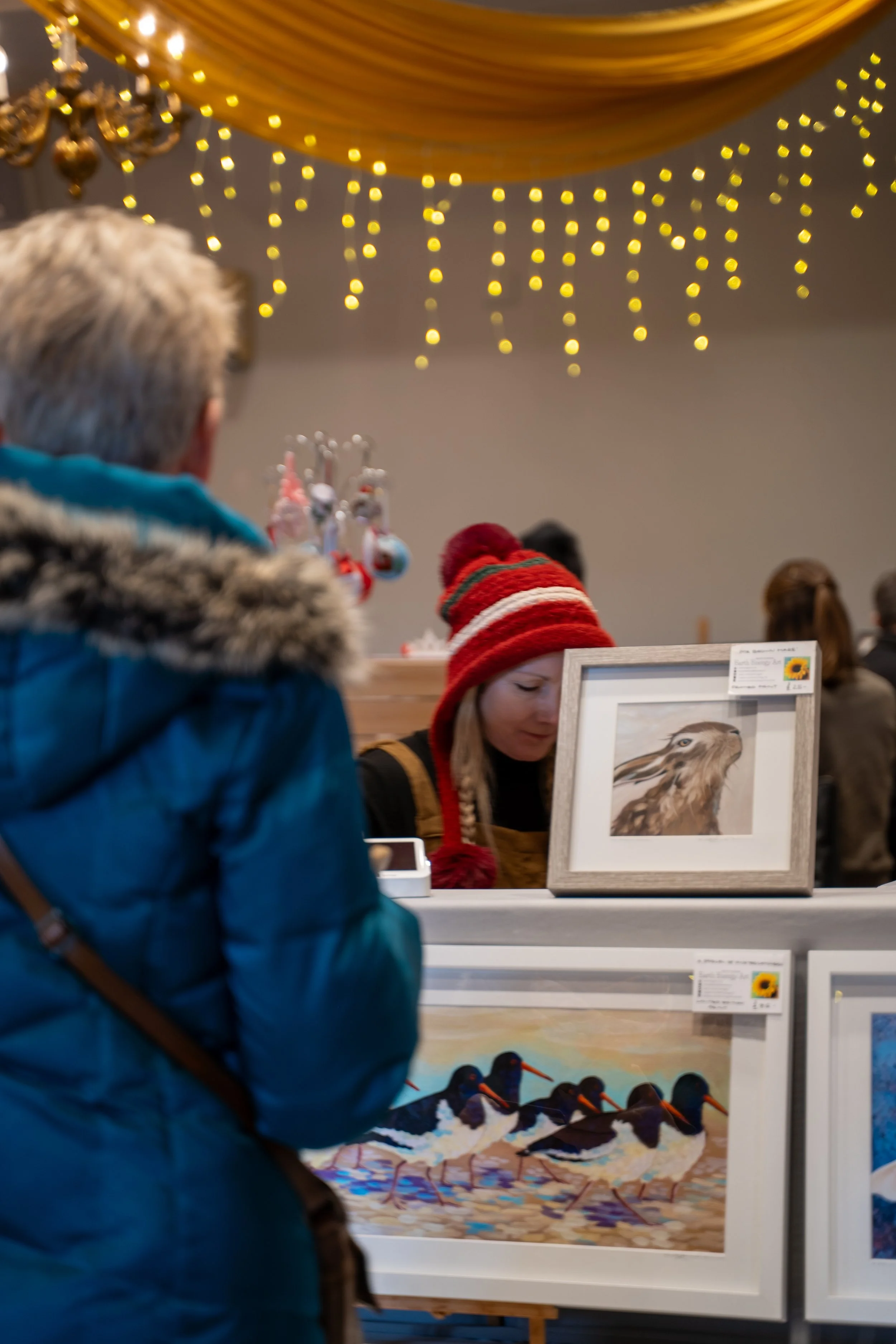 A person wearing a red and white striped winter hat looking at artwork of a brown owl with yellow eyes, behind a white display case, with other people in the background at an indoor art exhibition or gallery with yellow string lights on the ceiling.