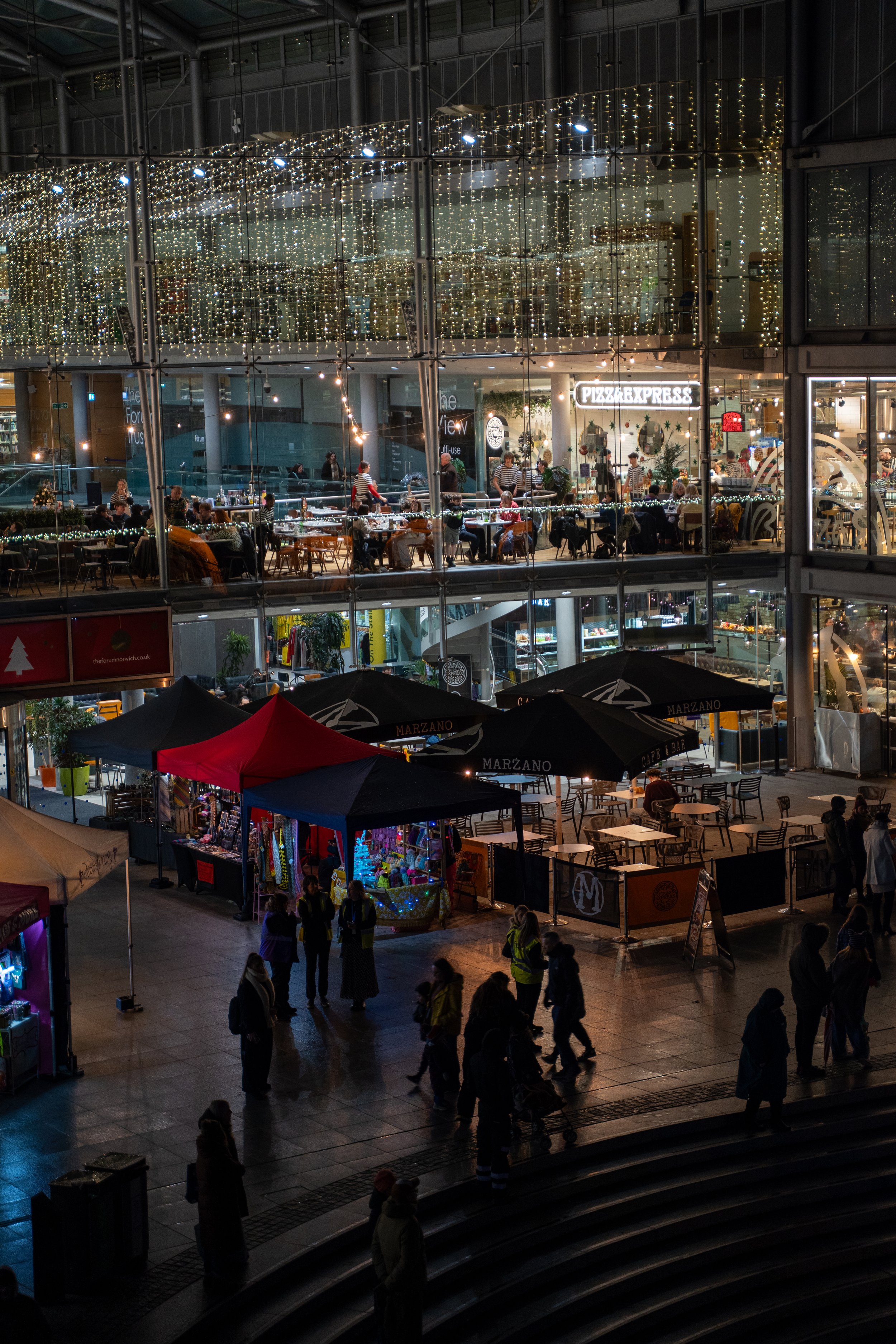 Night view of a multi-story shopping mall with bright decorative lights, people walking outside, and a dine-in restaurant called Pizza Express inside.