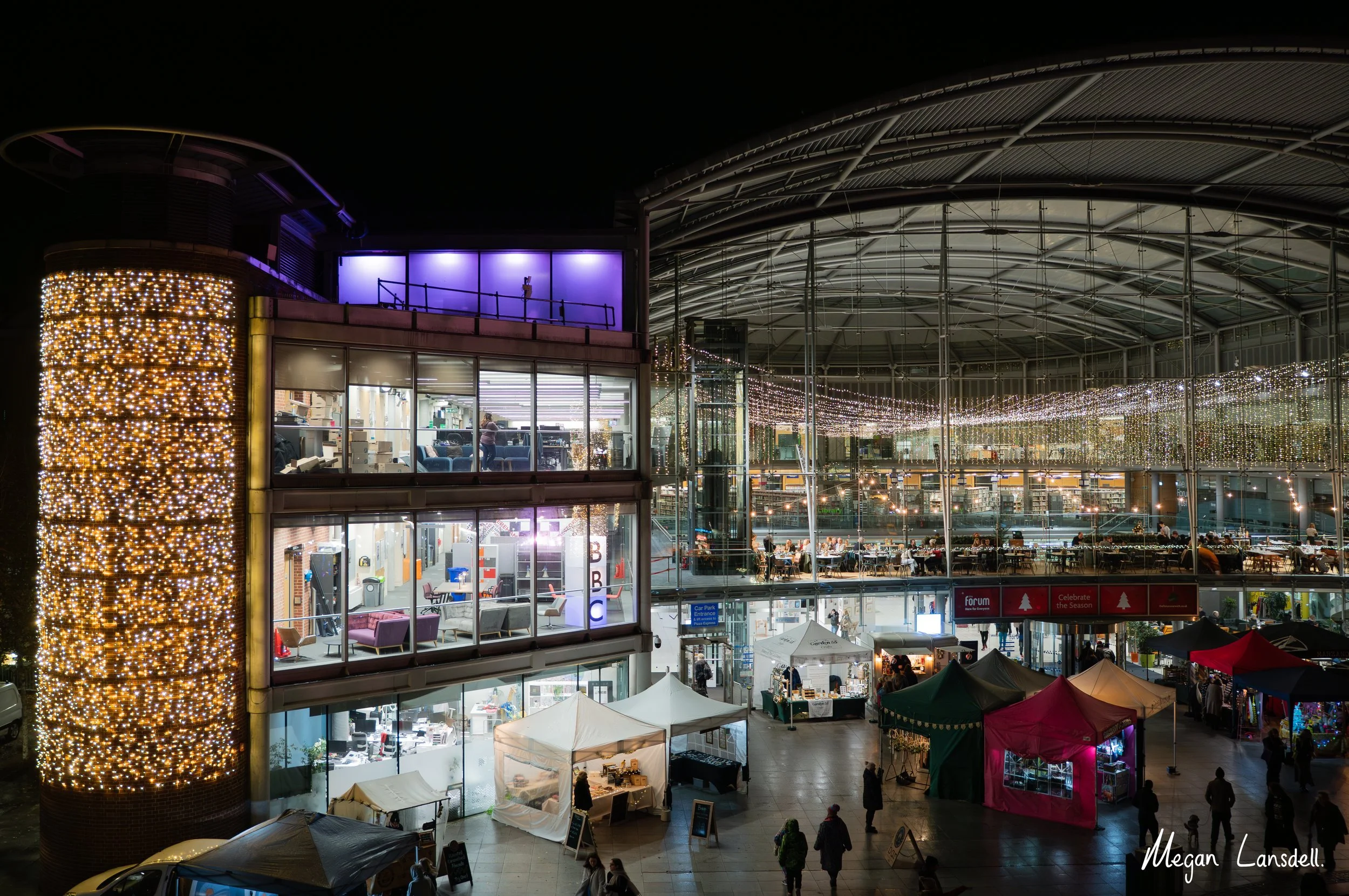 Night view of a modern, multi-level shopping mall with glass walls, decorated with string lights, and outdoor tents selling various goods at the entrance.
