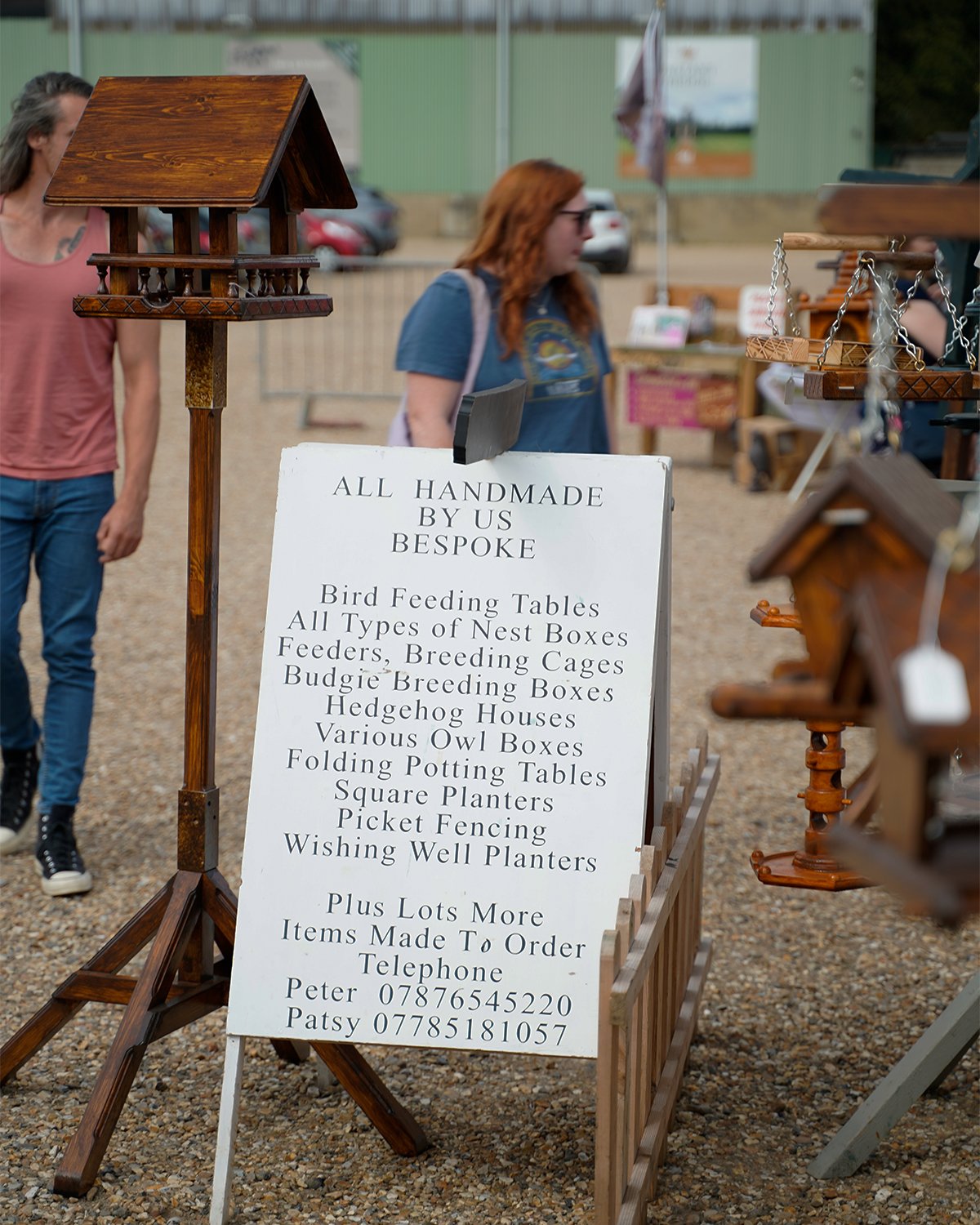 A sign at an outdoor market listing handmade birdhouses, nest boxes, feeders, cages, hedgehog houses, owl boxes, and planters, with contact information. Two people are visible in the background near wooden birdhouses and other crafts.