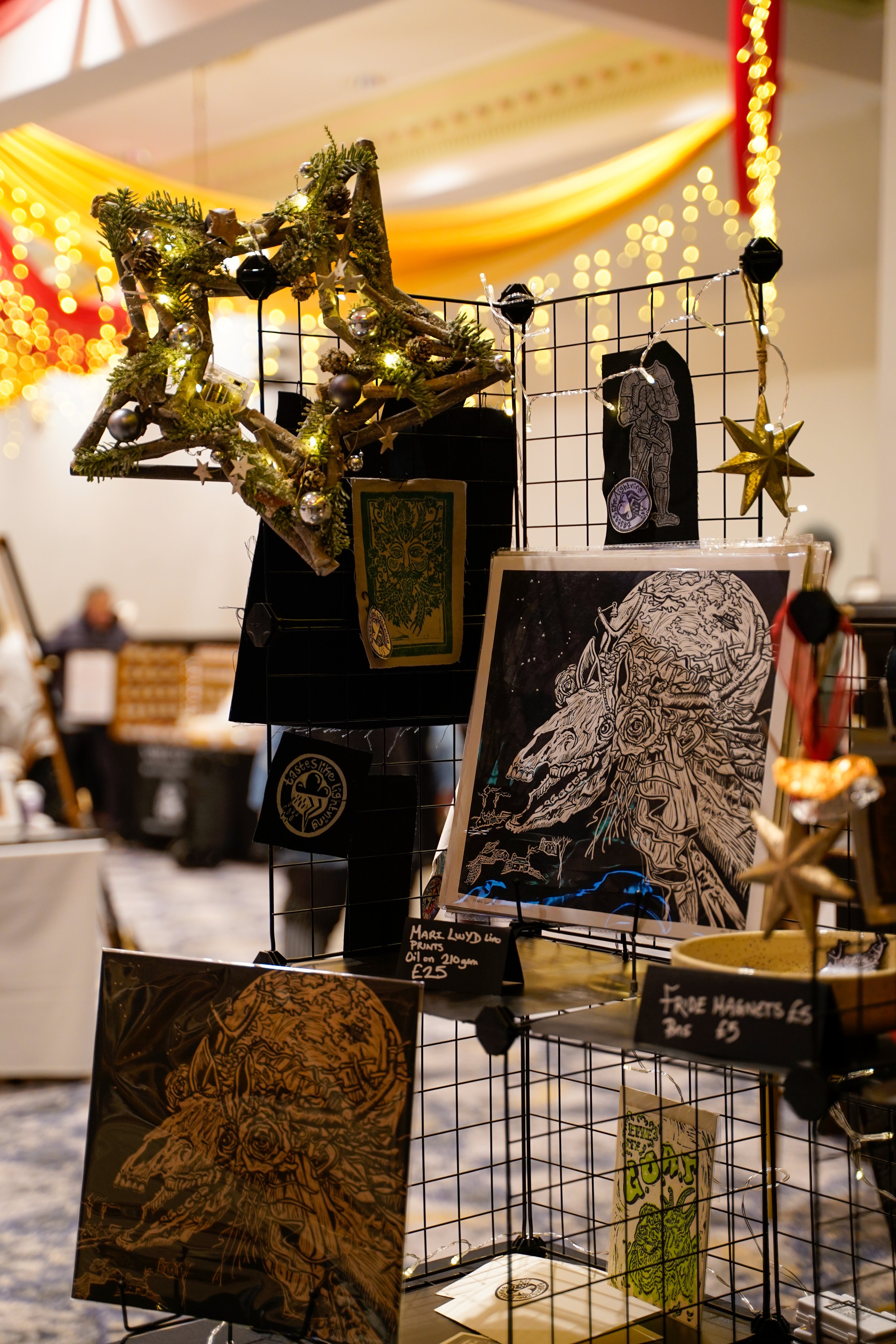 A display of art prints and greeting cards on a wire grid at an indoor market. The prints feature intricate black and white line drawings and colorful designs, with some text labels indicating prices.