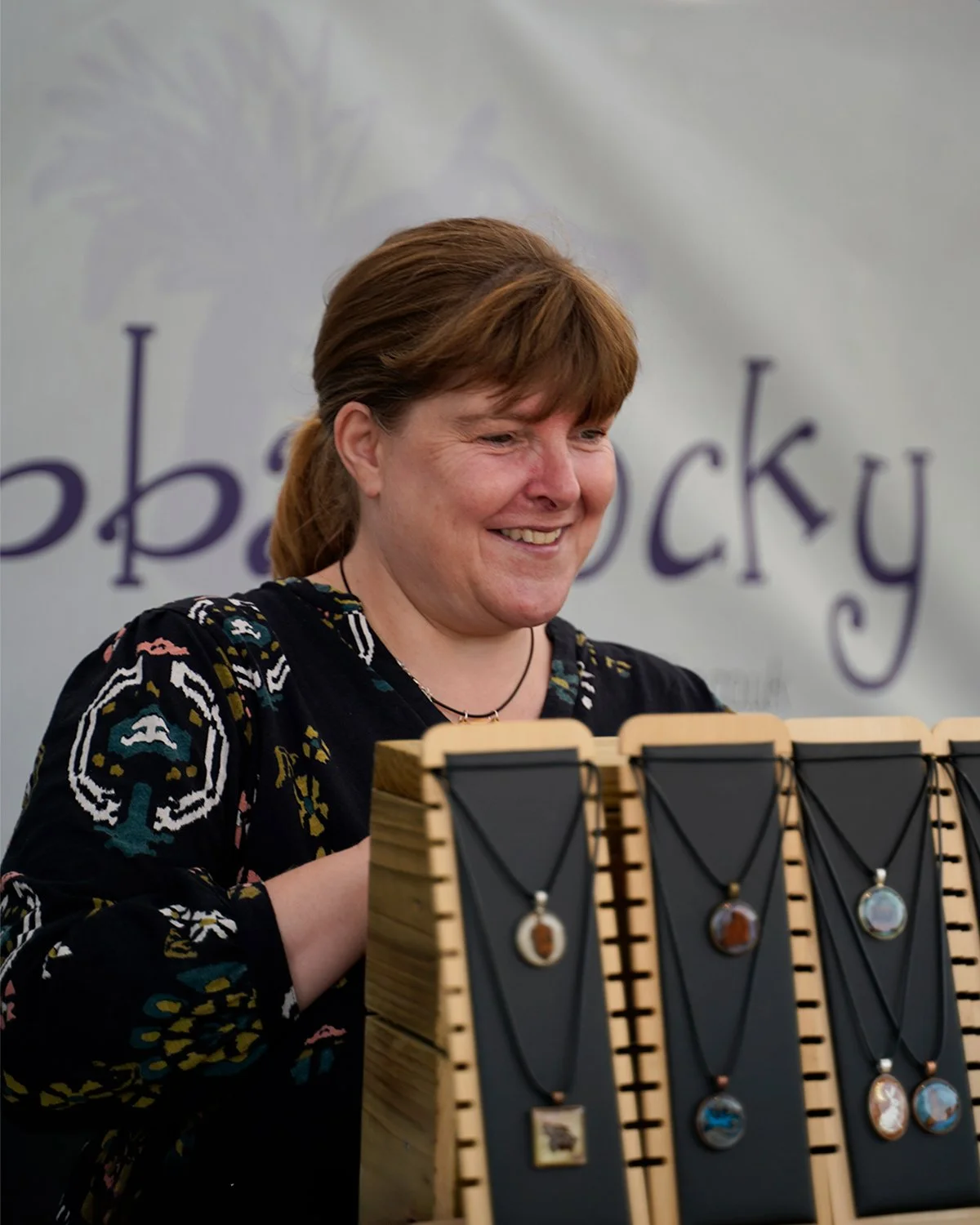 A woman with short, reddish hair smiling at a jewelry display featuring necklaces with round pendants, set on a wooden stand