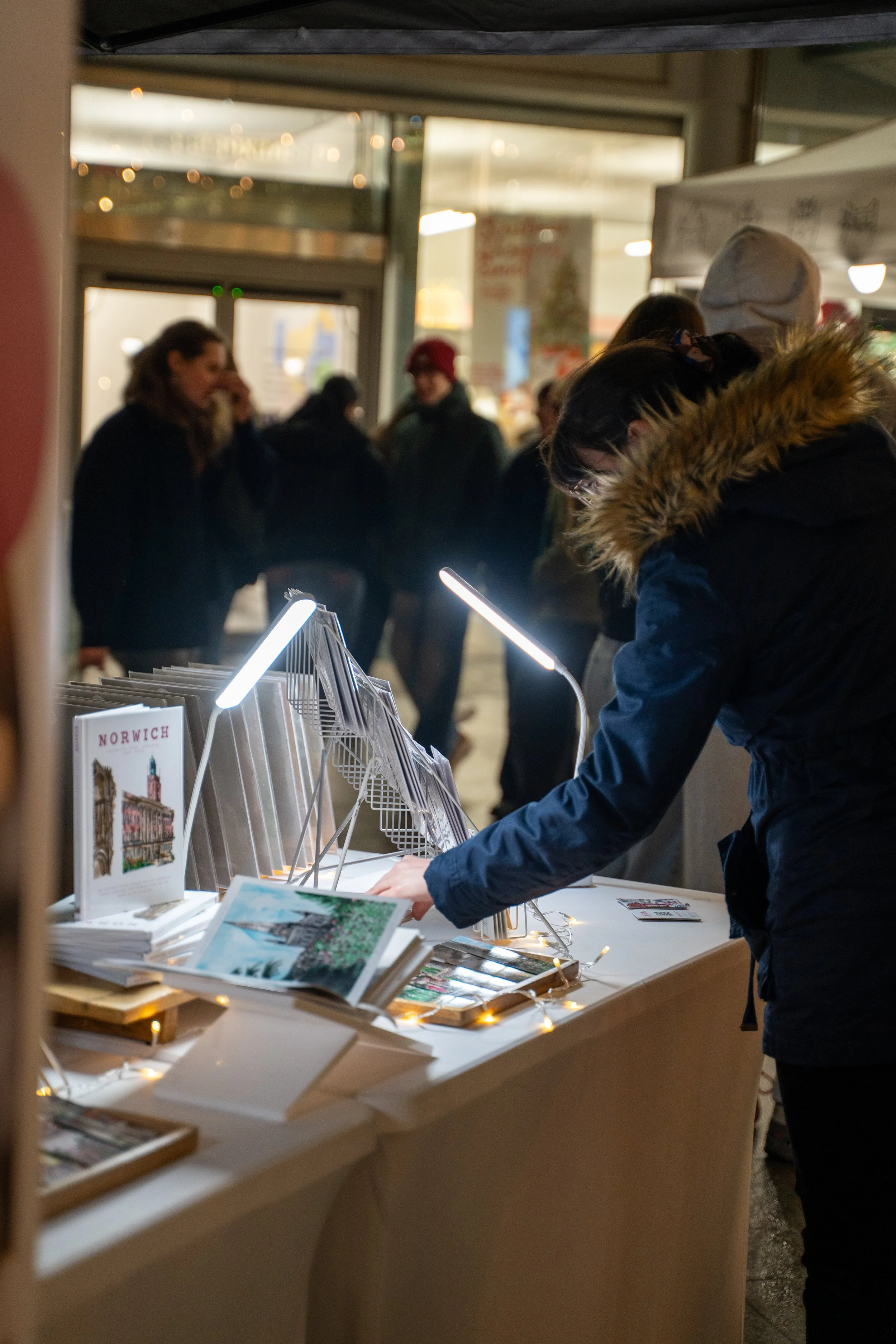 A person in a dark jacket with a fur-lined hood is browsing through postcards at an outdoor market stand decorated with string lights. Several people are visible in the background inside a building, some talking and looking at other displays.