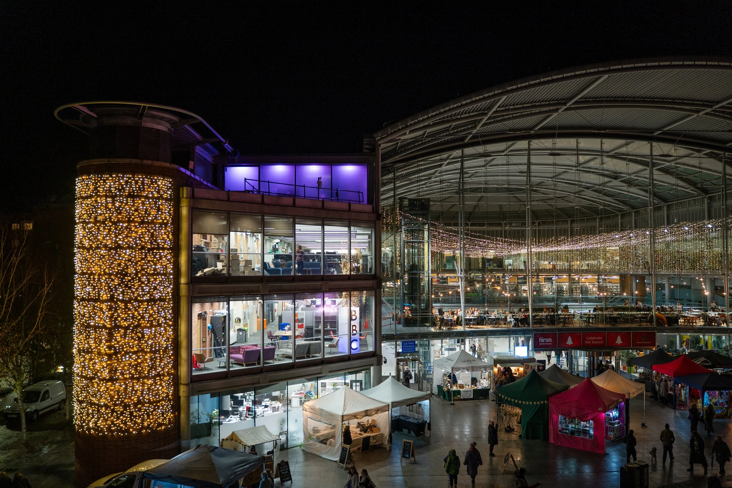 A multi-story building decorated with Christmas lights, with a market or event happening outdoors below, under a large metal canopy at night.