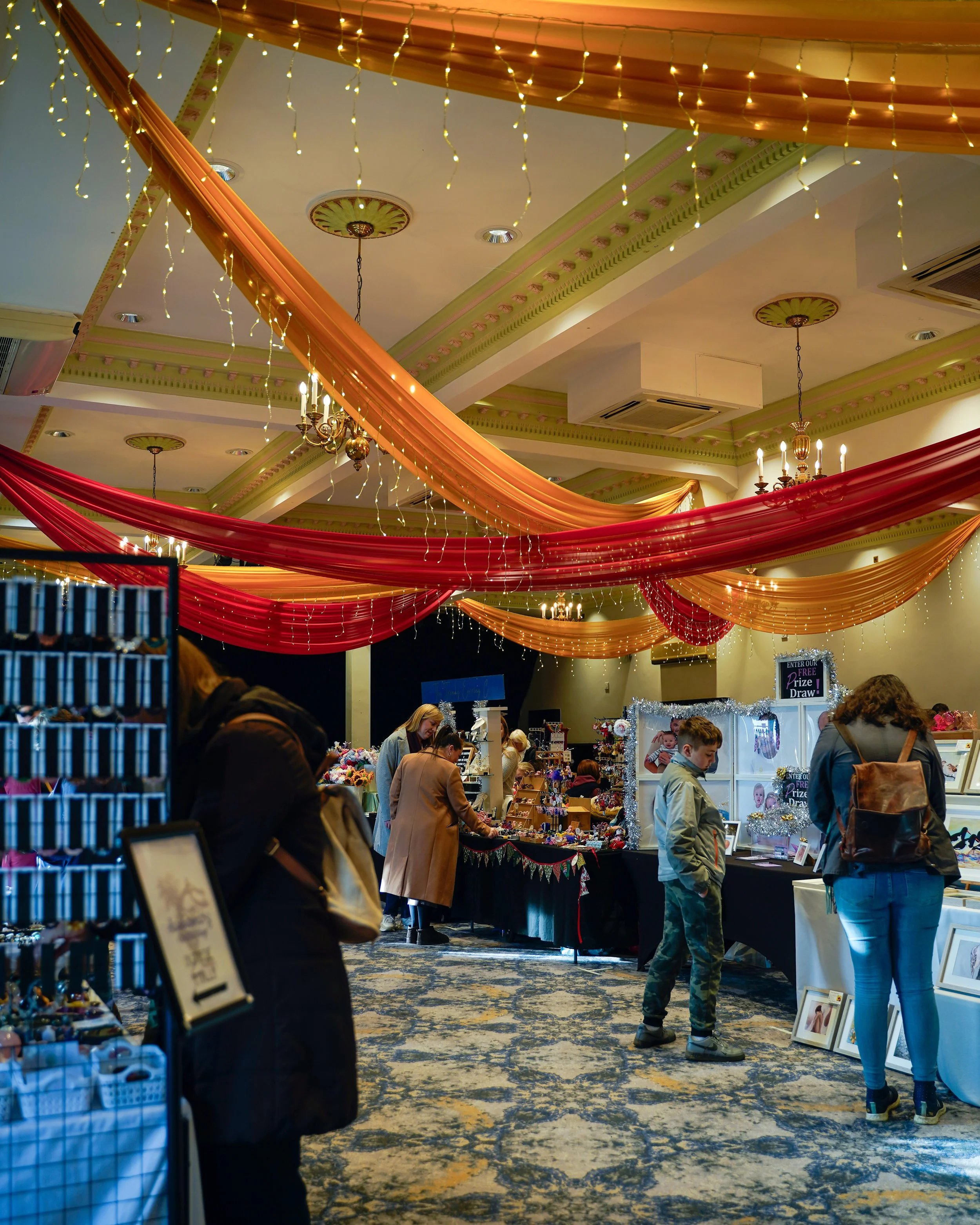 Indoor market or exhibition hall decorated with red and gold drapes and string lights hanging from the ceiling, with various vendors and shoppers browsing booths featuring jewelry, accessories, framed pictures, and other merchandise.