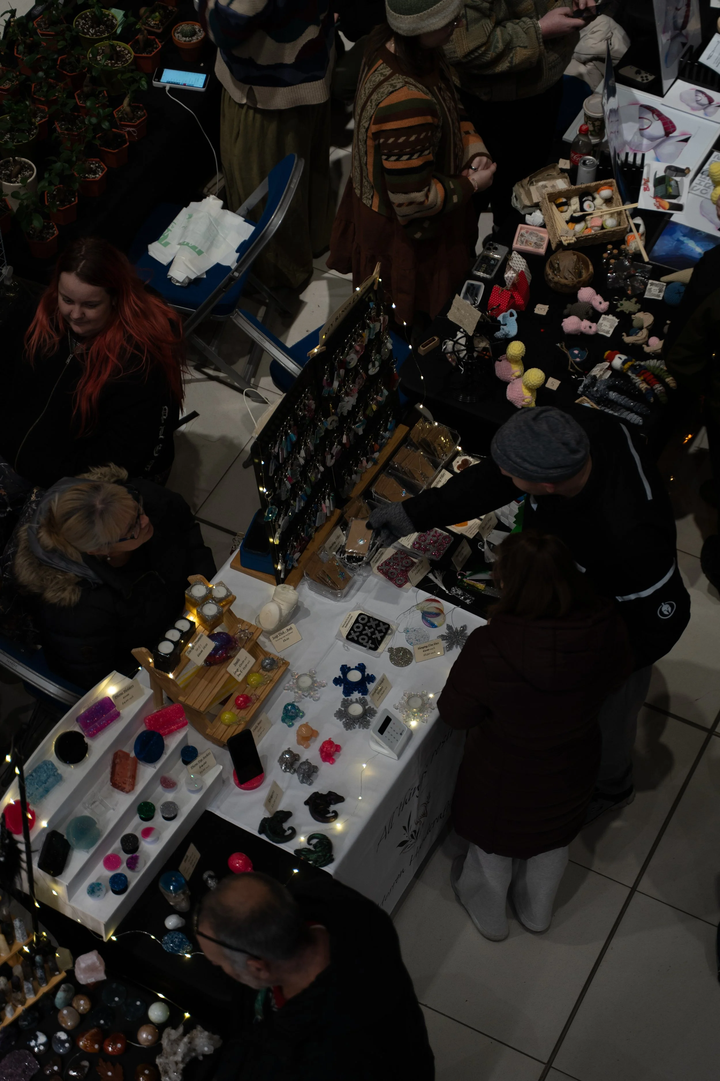 People shopping at a craft fair booth displaying various jewelry and decorative items, with some customers browsing and selecting items.
