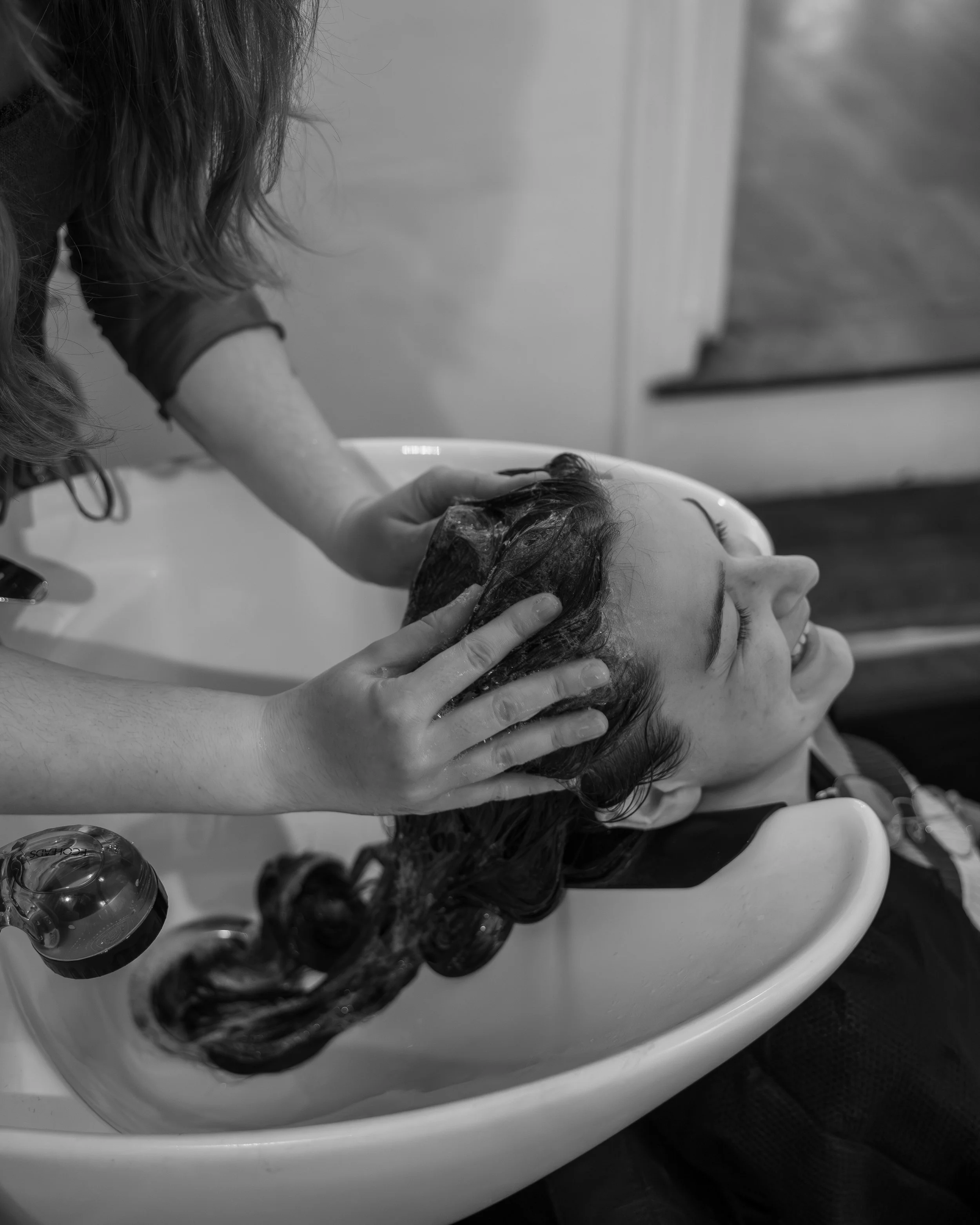 A person with wet hair leaning back in a salon sink while a stylist shampoos their hair.