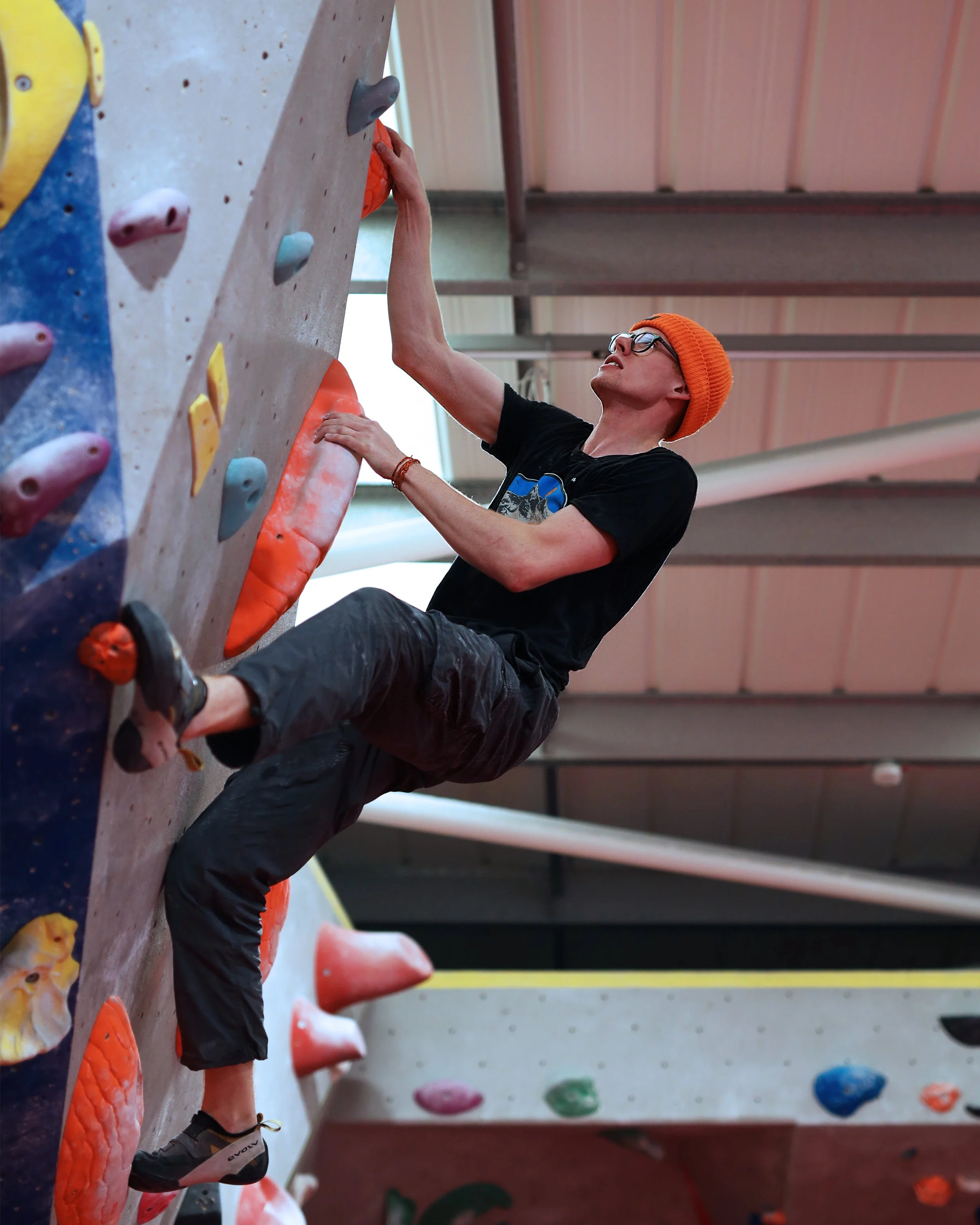 A person wearing glasses and an orange beanie is climbing an indoor rock wall with colorful holds. The person is wearing a black t-shirt and black pants, and is facing upward while gripping the holds.