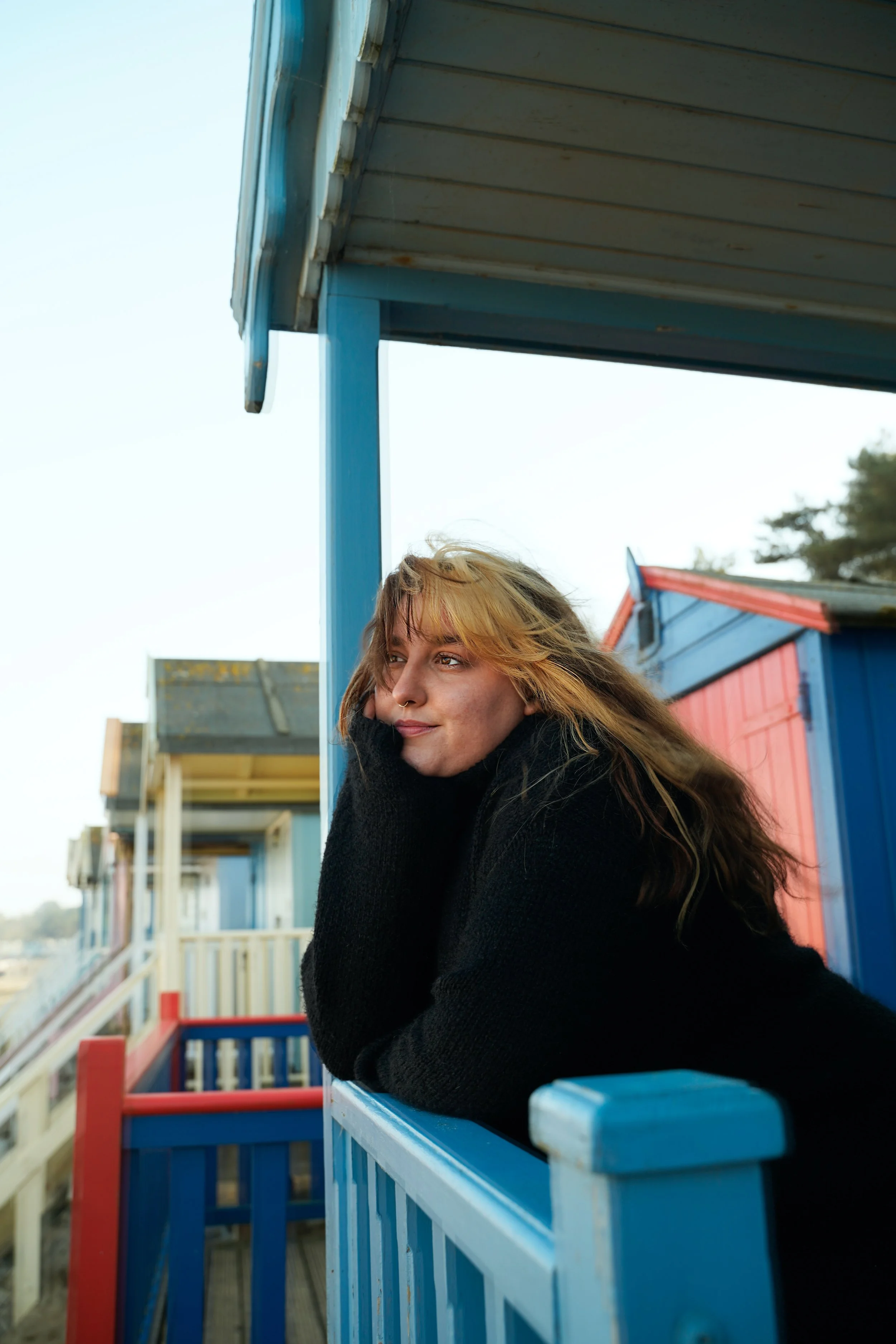 A woman with blonde hair and a nose piercing leaning on a blue railing outside a colorful beach hut, looking thoughtfully into the distance.