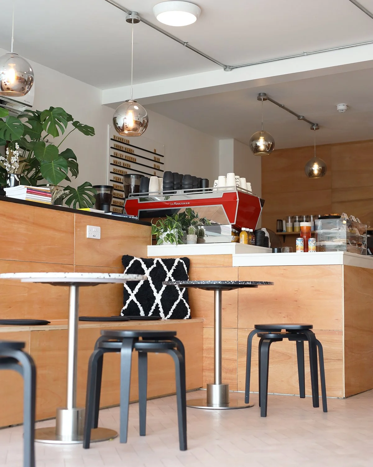 Interior of a modern coffee shop with wooden counters, black stools, a black and white pillow, a coffee machine, plants, and hanging lights.