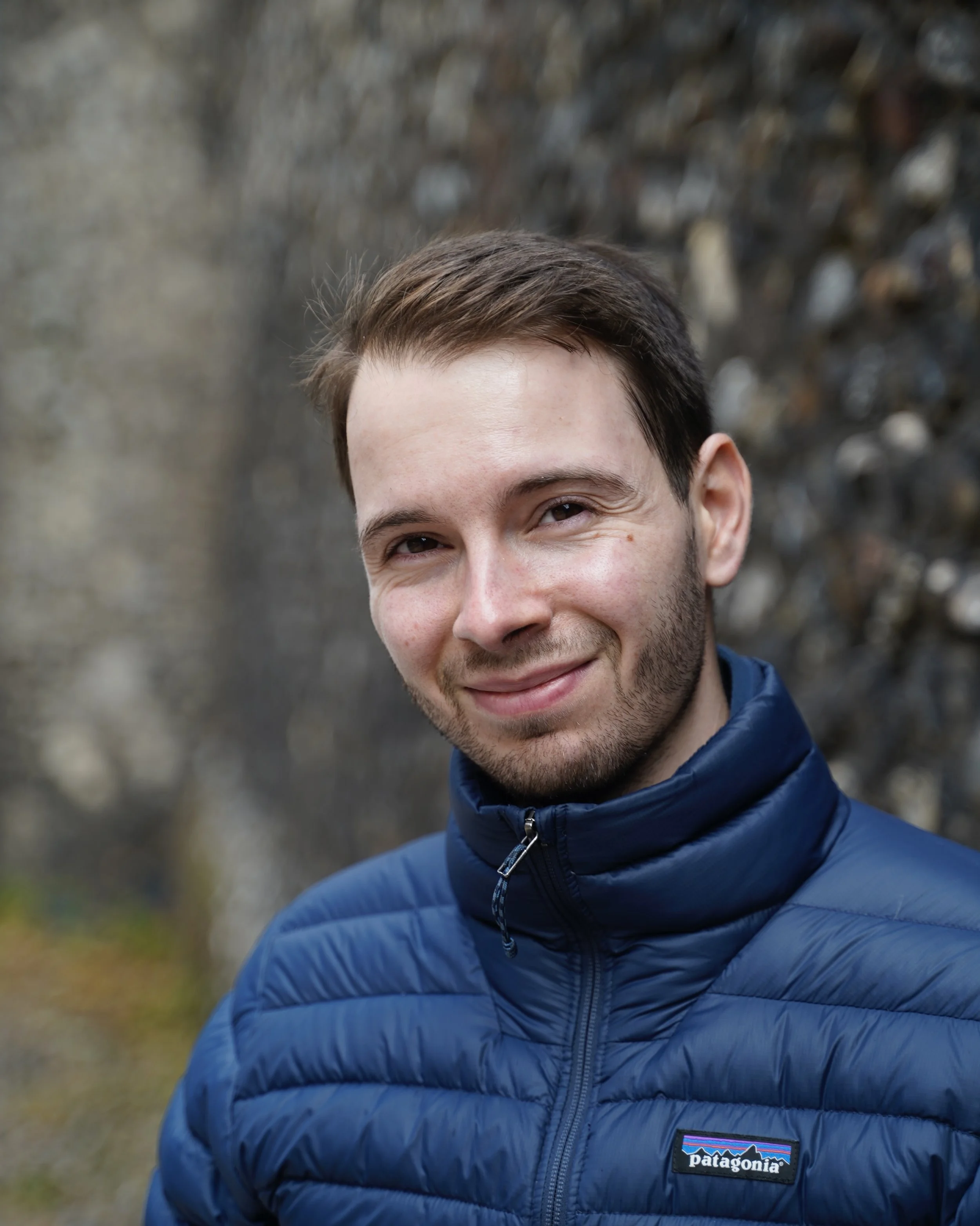 A smiling young man with brown hair and facial hair, wearing a blue Patagonia puffer jacket, outdoors with a blurred rocky and dirt background.