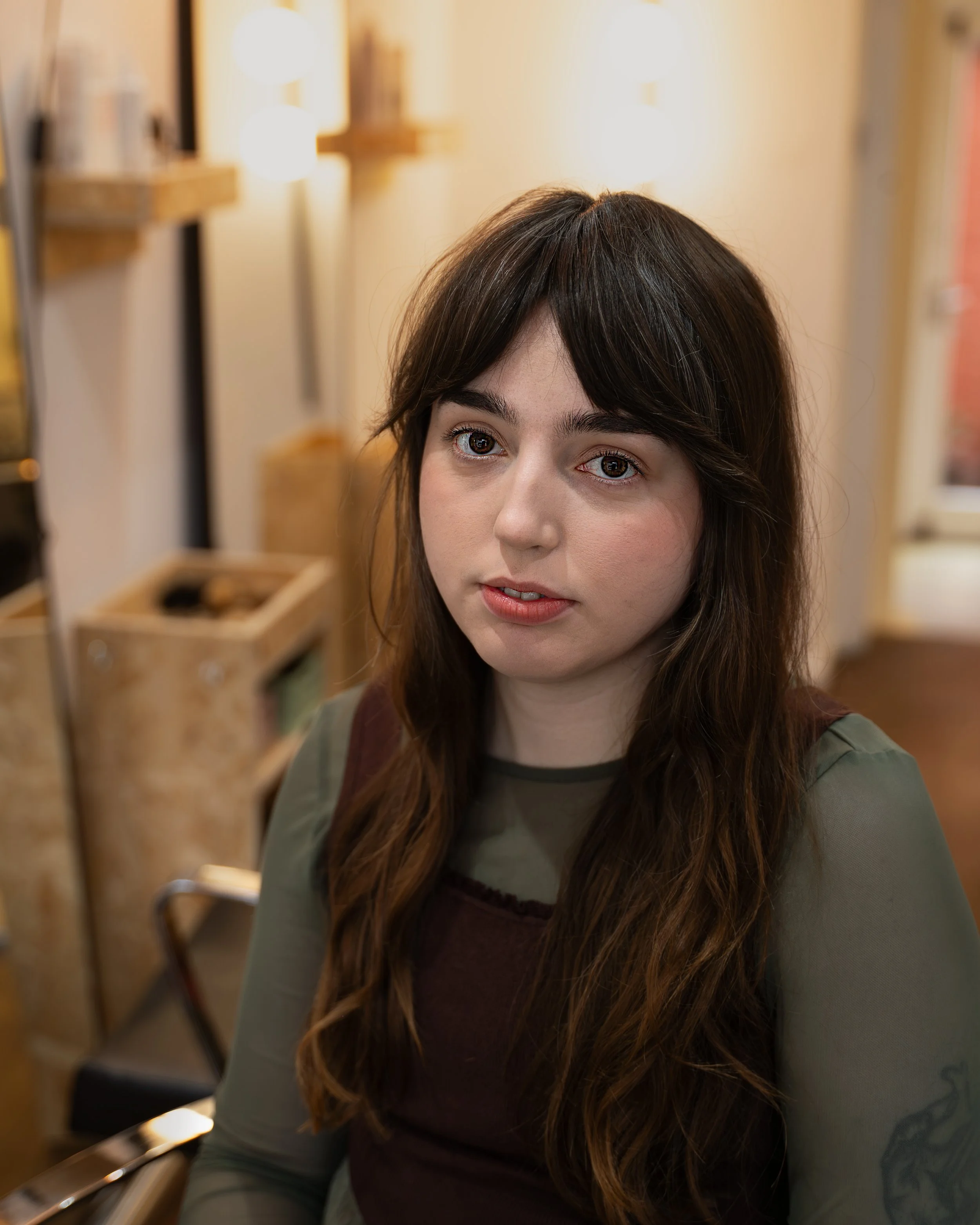 A young woman with long brown hair and light skin, sitting indoors near wooden shelves with lamps in the background, wearing a green shirt and a black top, looking slightly to her left.
