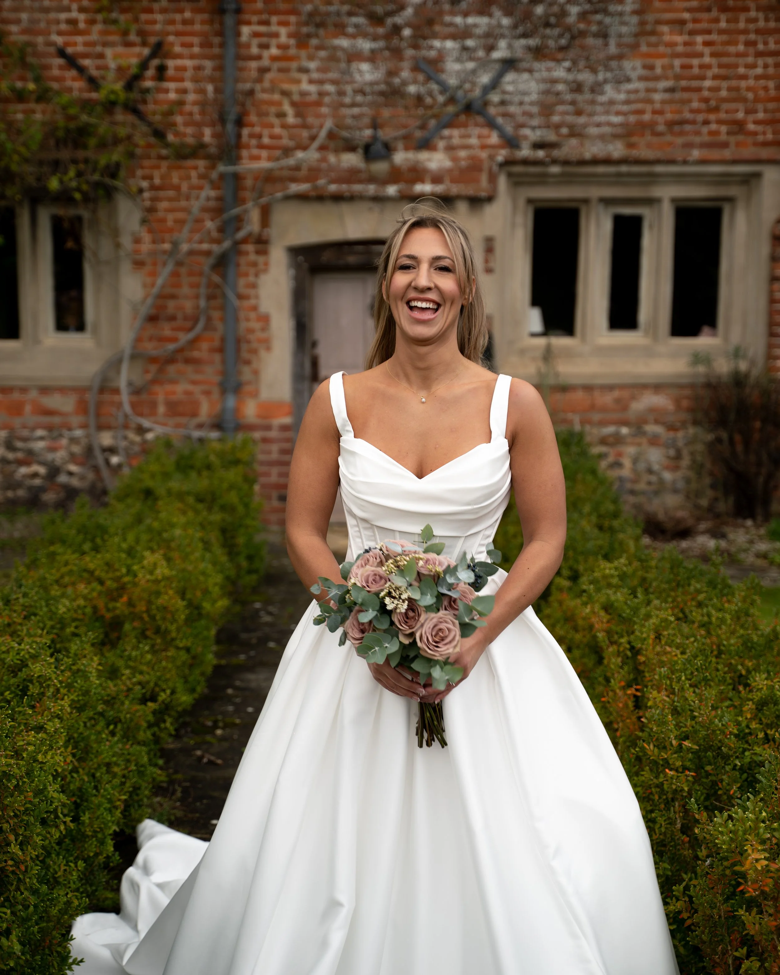A smiling woman in a white wedding dress holding a bouquet of flowers, standing outdoors in front of a brick building with windows and green bushes.