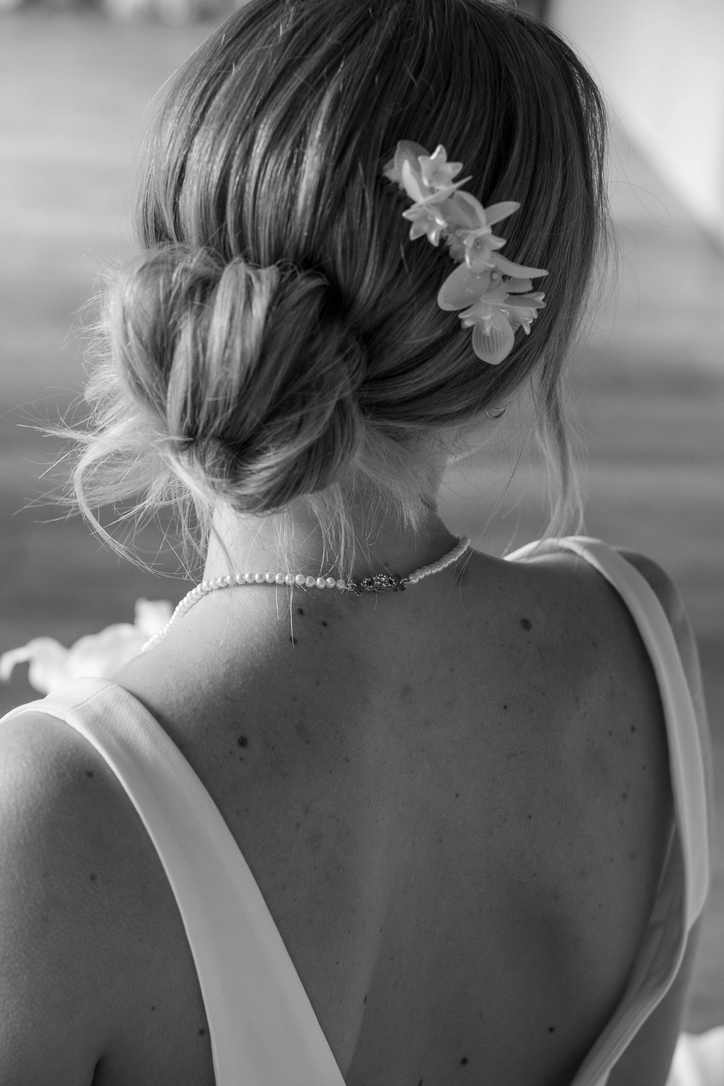 A woman with a braided hairstyle adorned with flowers, wearing a sleeveless top and a pearl necklace, seen from behind in black and white.