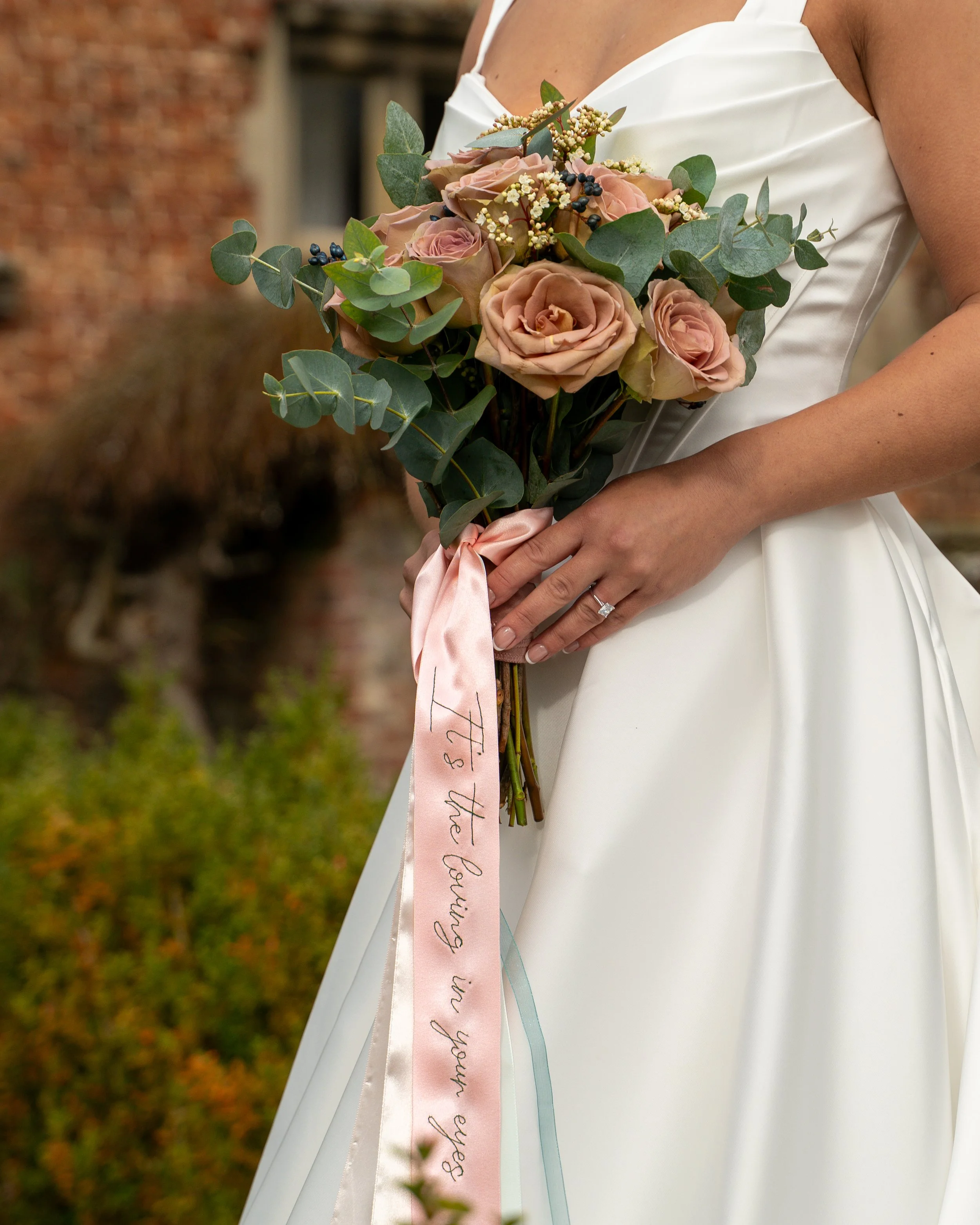 Close-up of a bride holding a bouquet of pink roses, greenery, and small white flowers, tied with a pink ribbon with cursive writing, in front of an outdoor rustic background.
