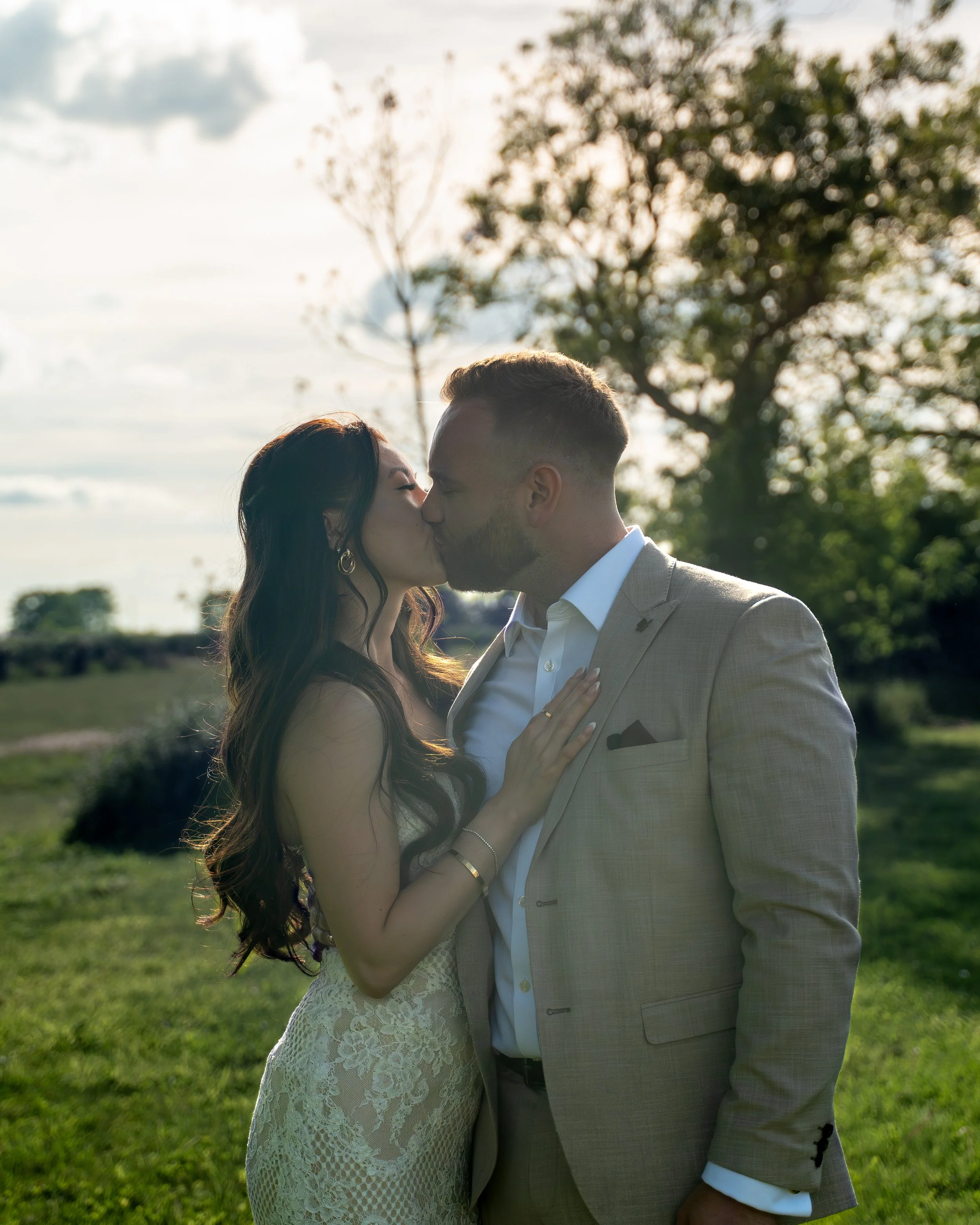 A couple, dressed in wedding attire, sharing a kiss outdoors during daytime with trees and cloudy sky in the background.