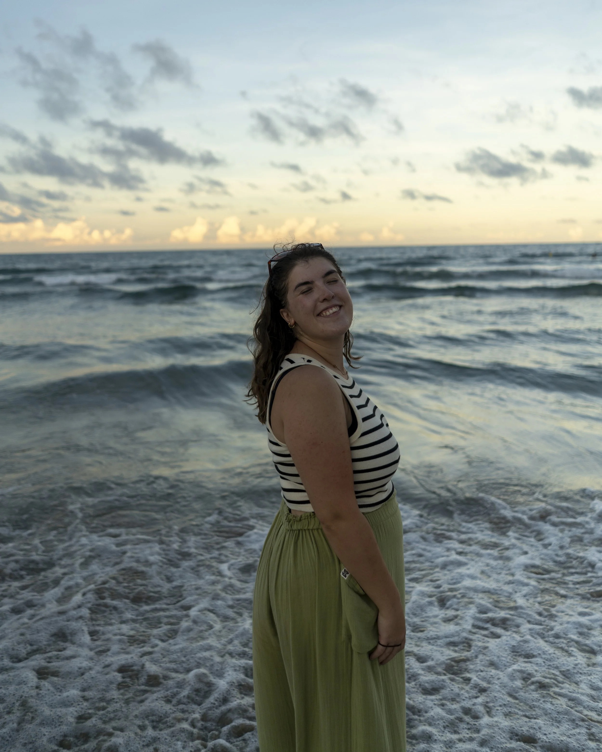 A young woman with dark hair smiling with eyes closed, standing at the beach with ocean waves in the background during sunset.