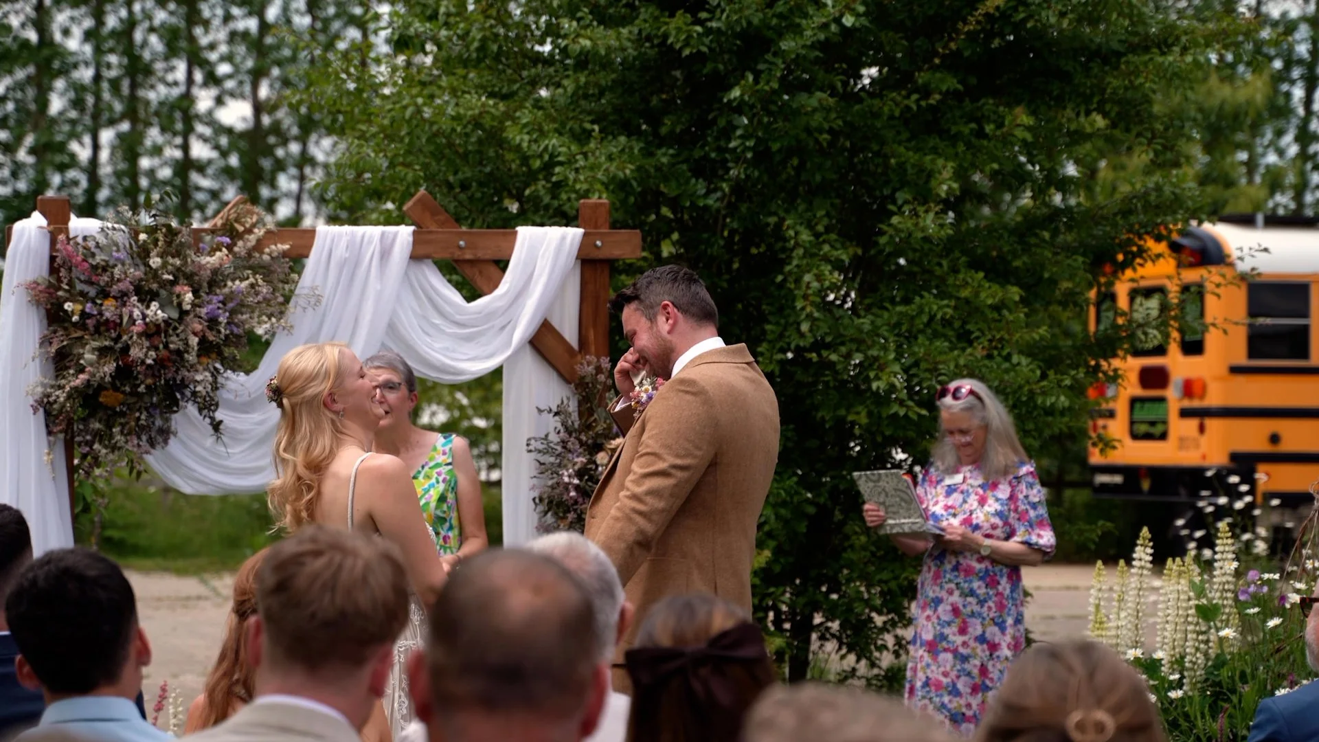 A bride and groom exchange vows at an outdoor wedding ceremony beneath a wooden arch decorated with white fabric and flowers, with guests seated in front, and a woman reading from a book in the background.