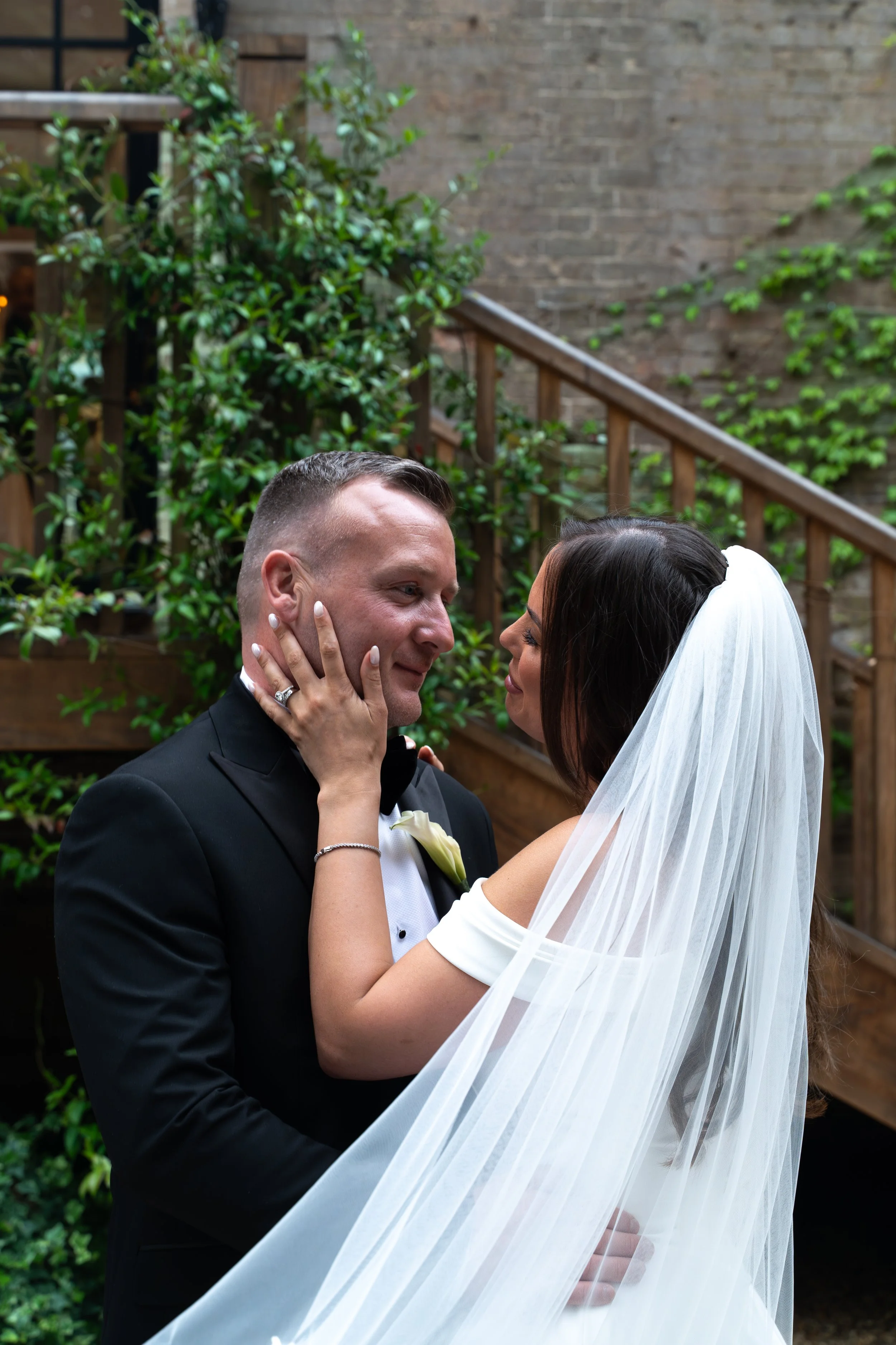 A bride and groom are holding each other and smiling during their wedding in an outdoor setting with wood stairs, railing, and greenery.