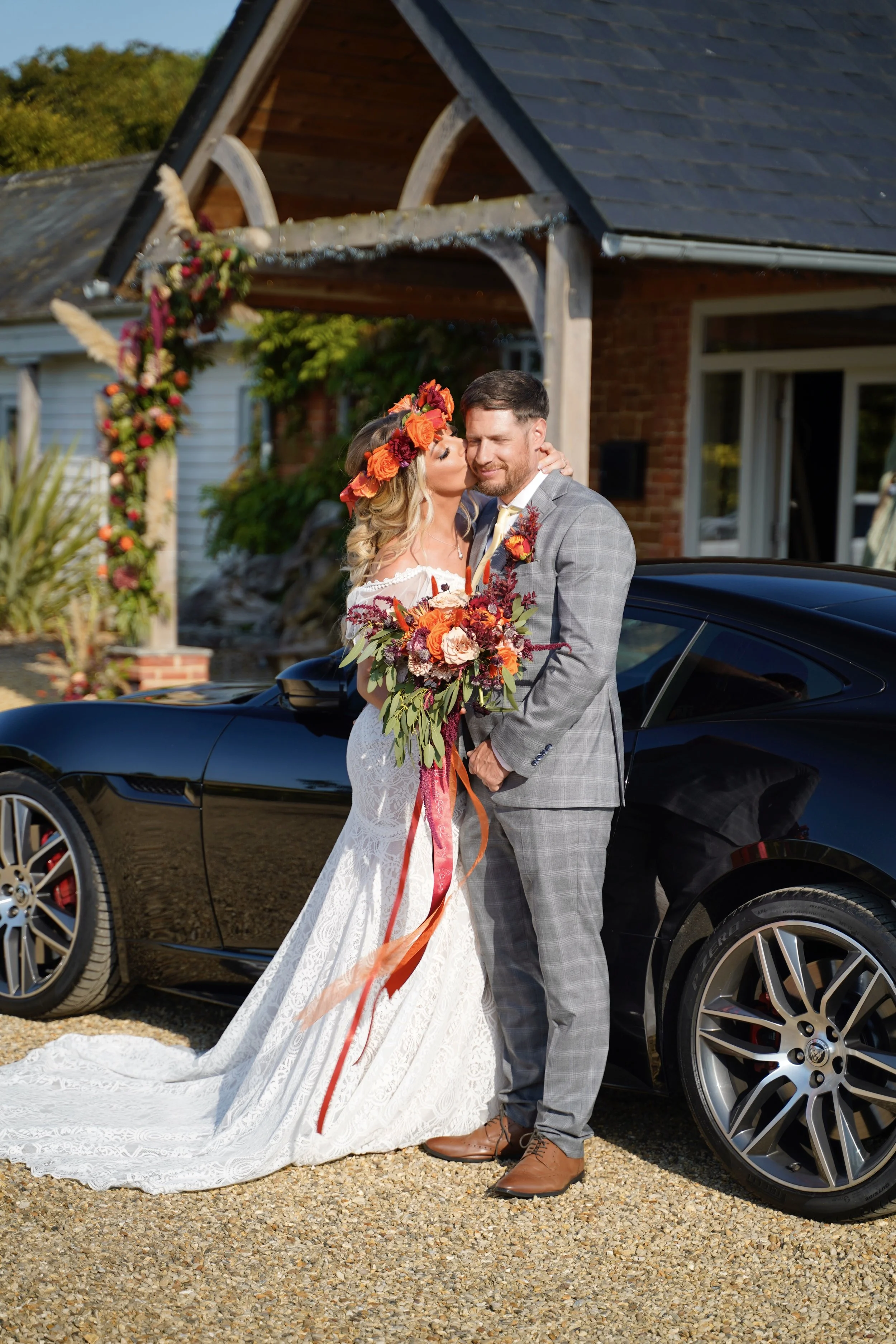A bride and groom embrace in front of a black sports car outside a house decorated with flowers for a wedding.