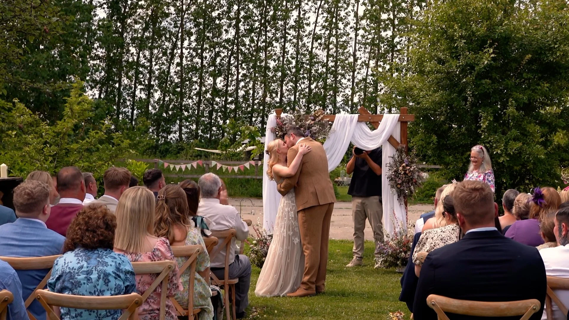 A couple shares a kiss during a wedding ceremony outdoors, with guests seated around them and a decorated arch in the background.