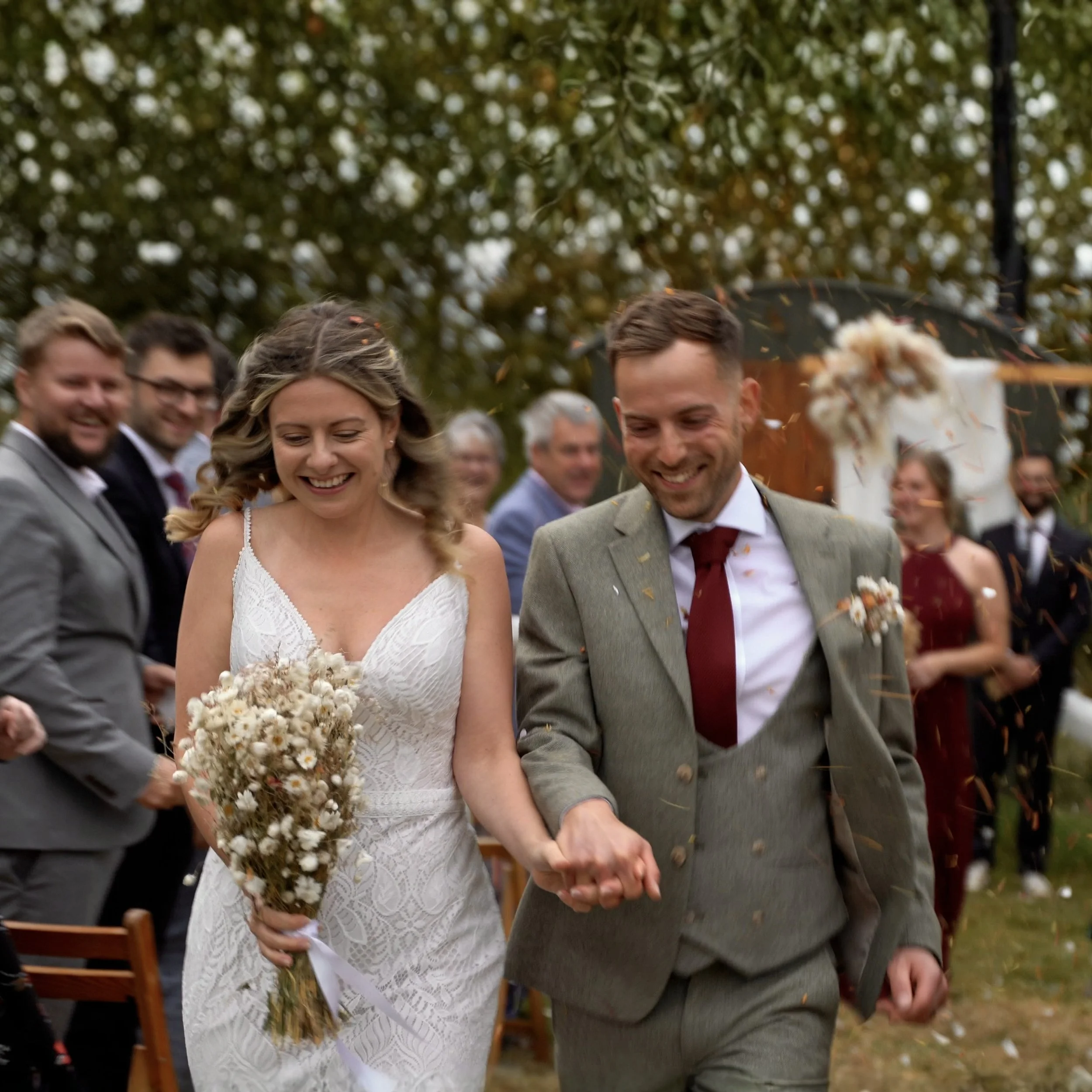 Bride and groom holding hands during an outdoor wedding ceremony, with guests seated and smiling in the background, autumn trees, and falling autumn leaves.