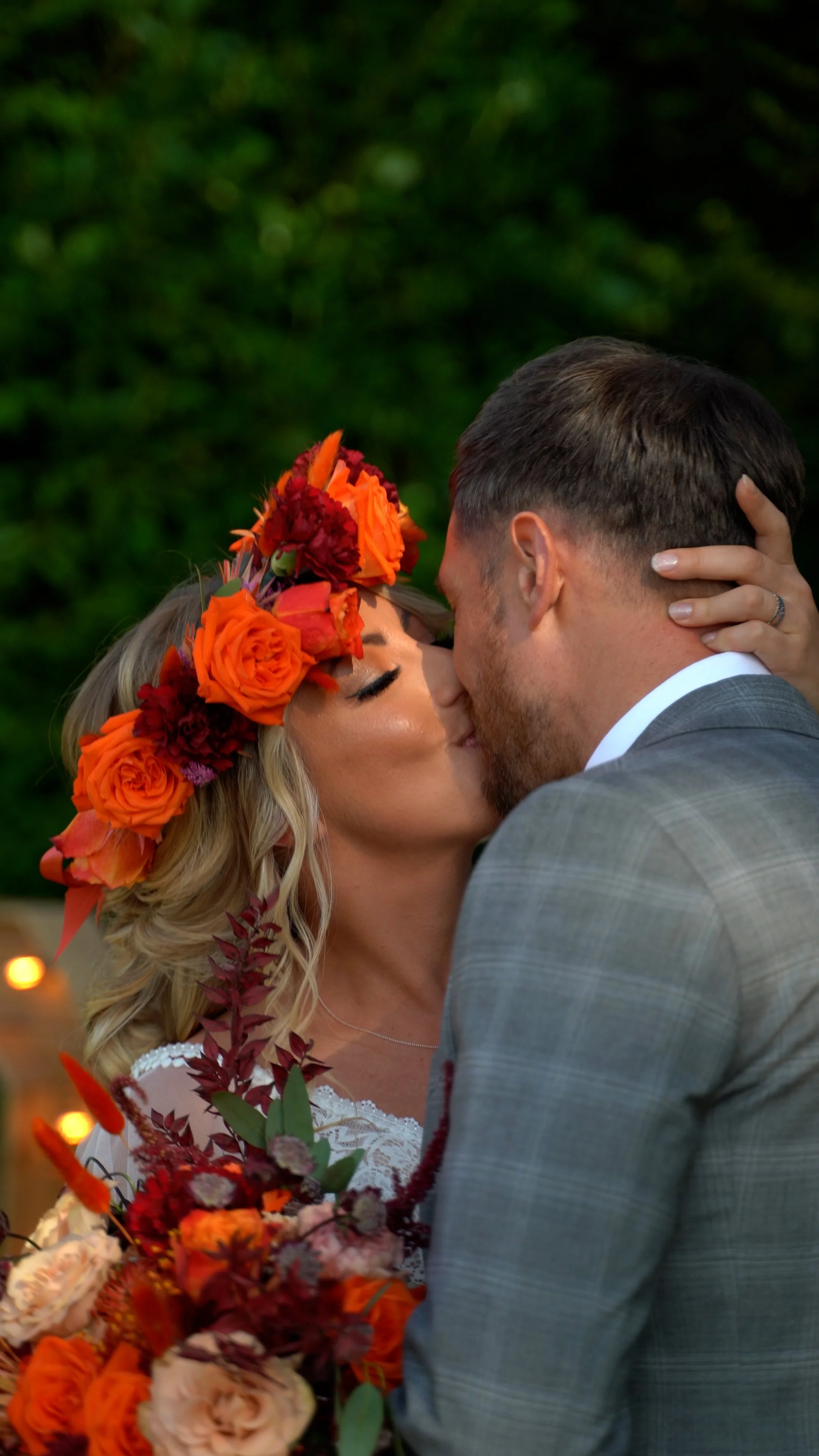 A couple kissing at a wedding, with the woman wearing a floral crown and holding a bouquet of flowers.