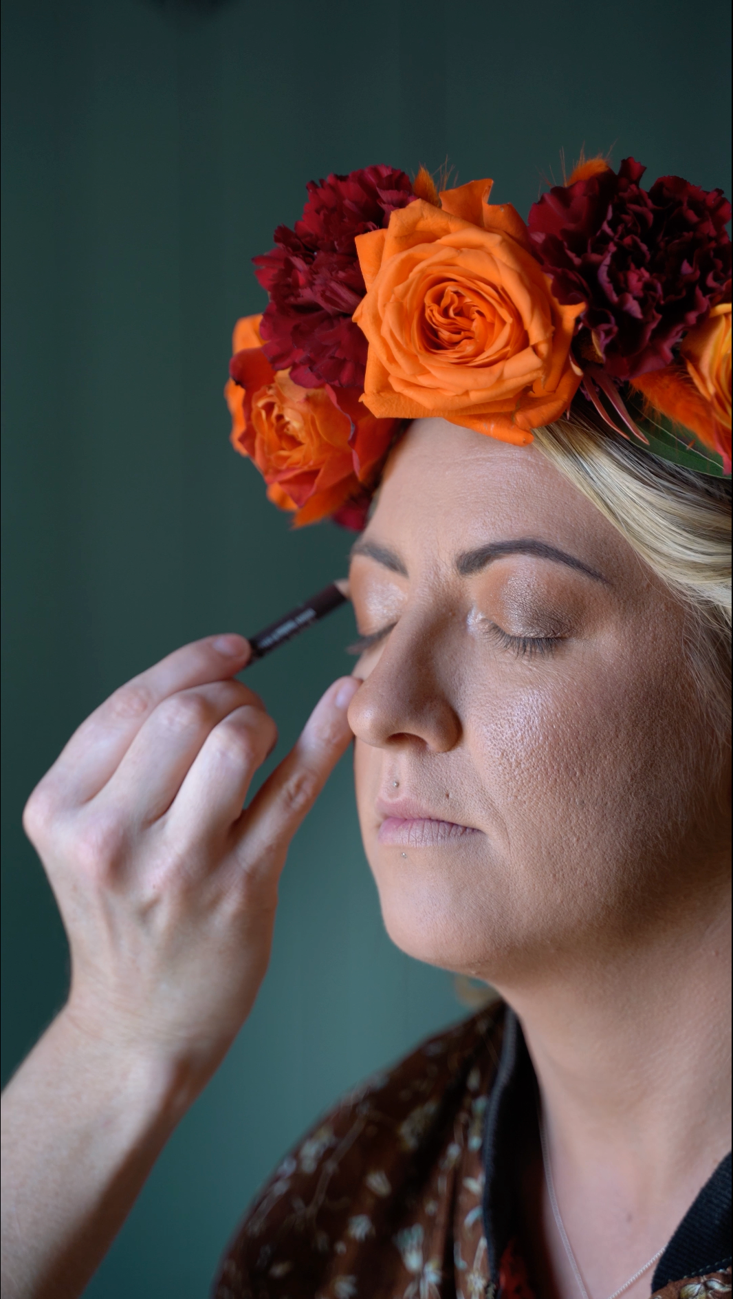 A woman with a floral crown is getting her makeup done, with her eyes closed.
