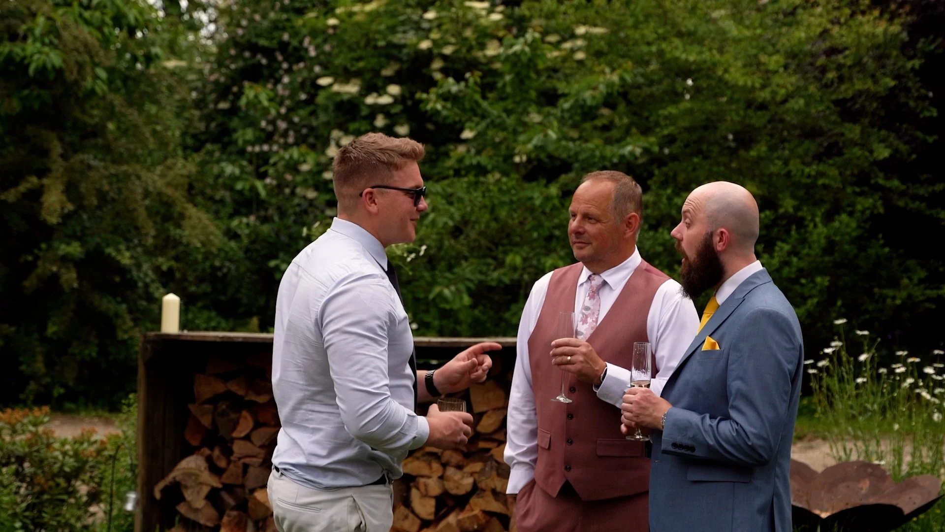 Three men in formal attire having a conversation outdoors at a social event, two holding champagne glasses, with lush greenery in the background.