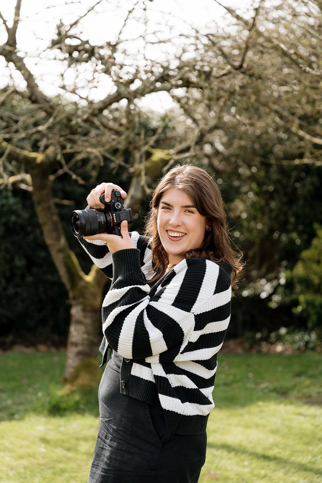 A woman with brown hair smiling and holding a camera outdoors near a tree.