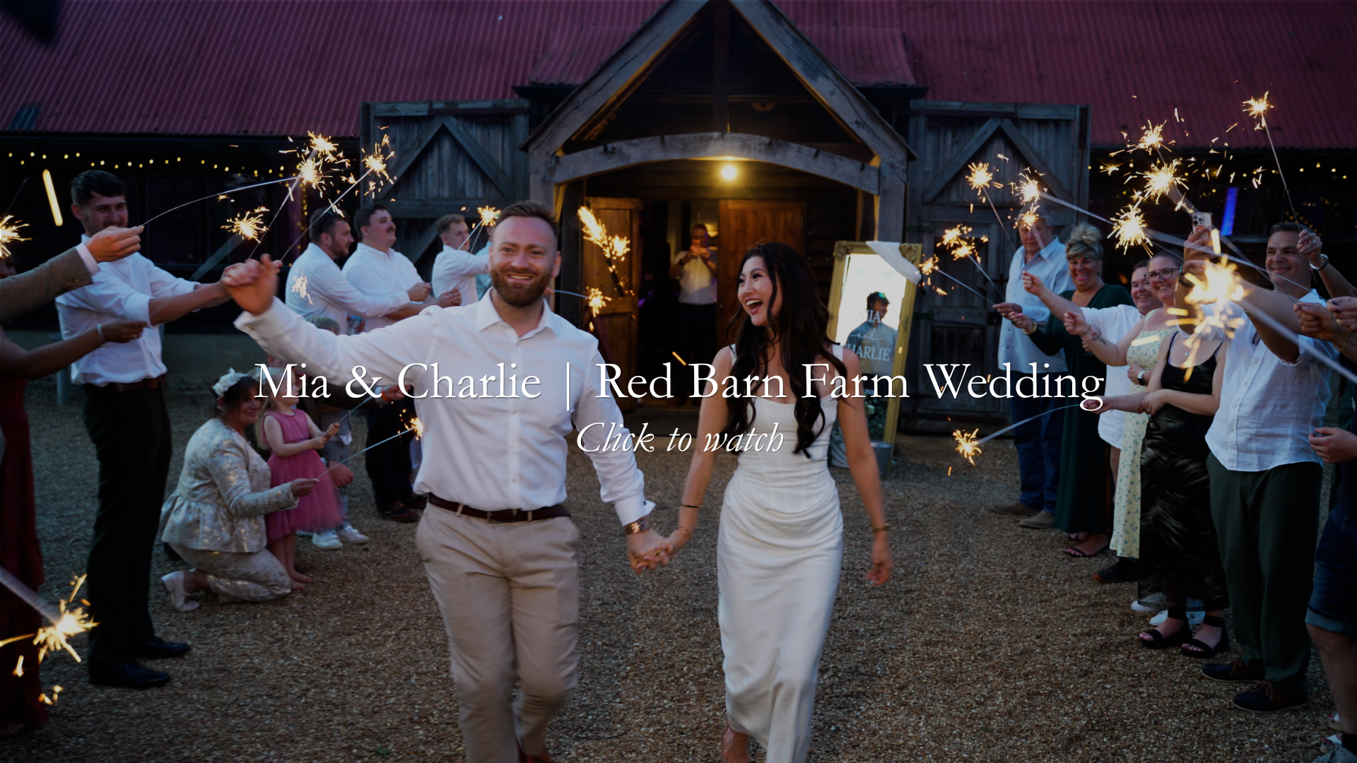 A newlywed couple holding hands and walking through a celebration with sparklers, surrounded by friends and family outside a rustic barn during a wedding at night.