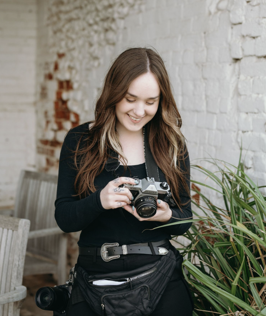 A young woman with long brown hair, smiling and looking at a camera she is holding, standing outdoors near a white brick wall and green plants.