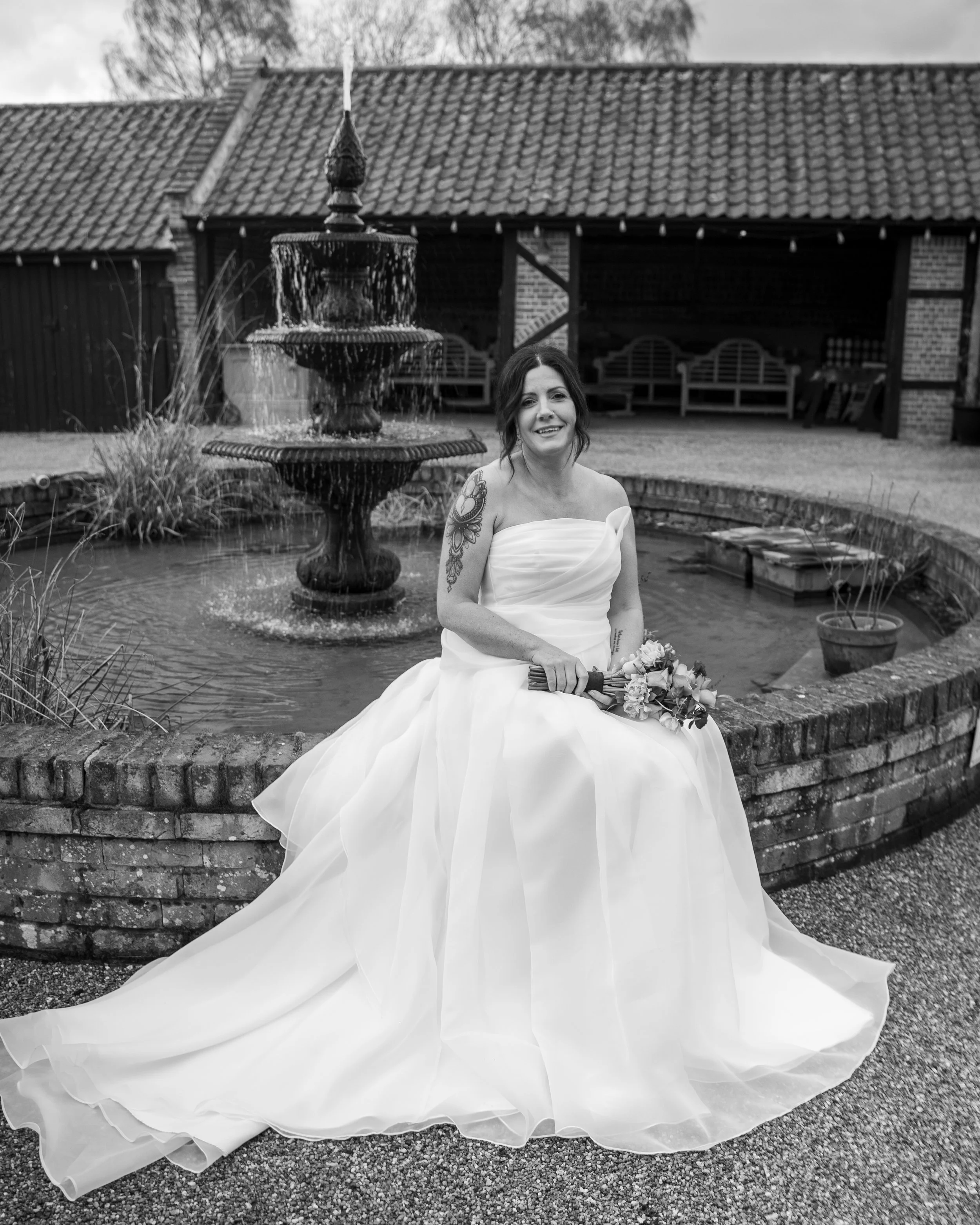A woman in a wedding dress sitting on a brick ledge in front of a fountain, holding a bouquet, with a building and trees in the background.
