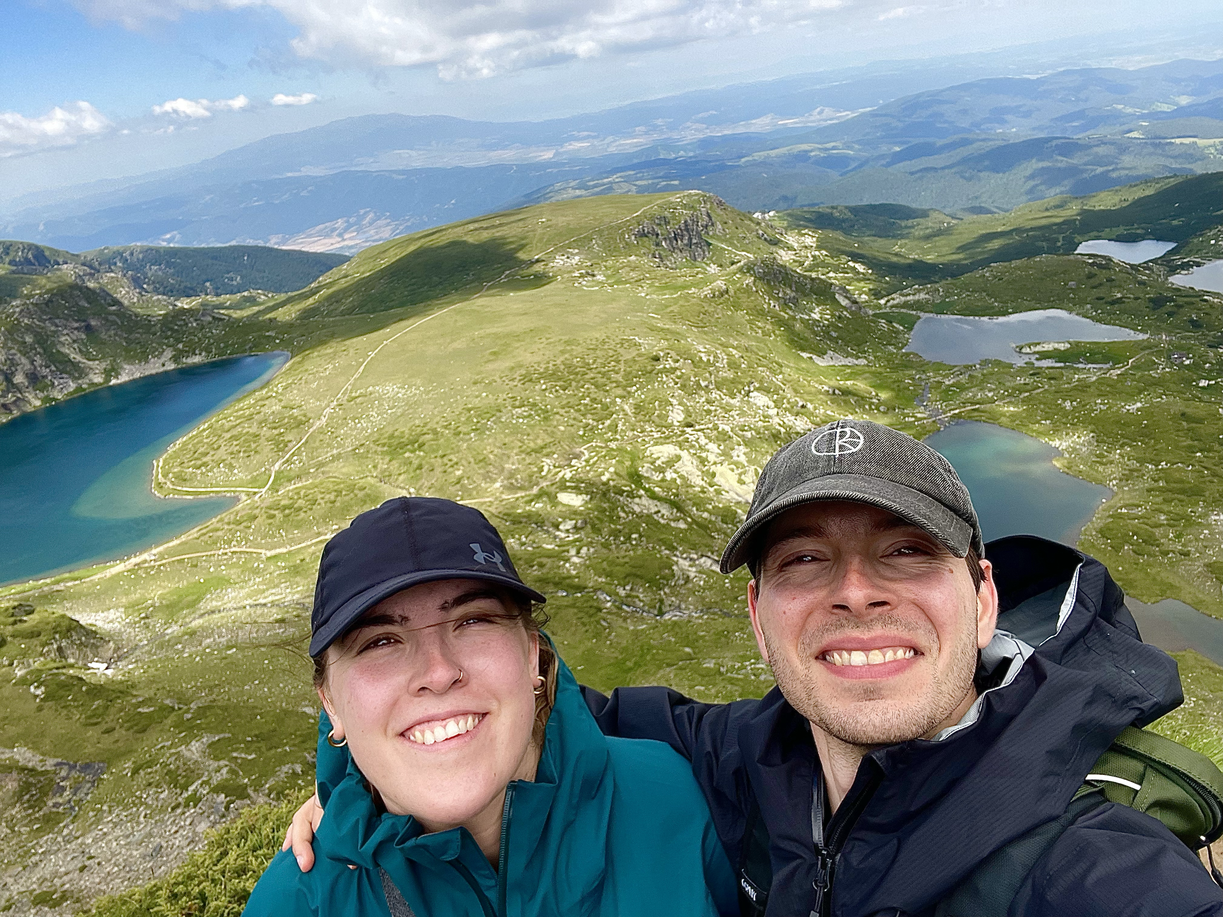 Two smiling hikers, a woman with short hair, earrings, and a nose ring, and a man with short hair, are taking a selfie on a mountain trail. Behind them is a scenic view of rolling green hills, mountain peaks, and lakes under a partly cloudy sky.