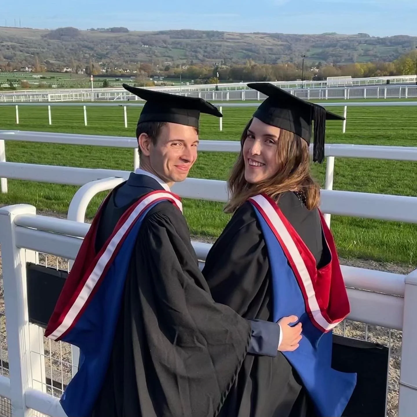 Two students in graduation gowns and caps sitting on a white fence at a racecourse, smiling at the camera with a scenic hilly landscape in the background.