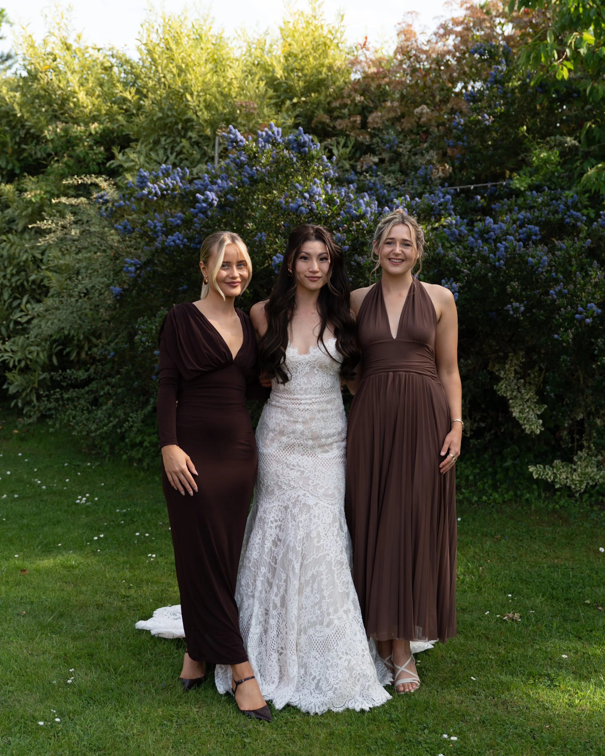 Three women in elegant dresses standing outdoors on grass, with lush green trees and blue flowers in the background, smiling at the camera.