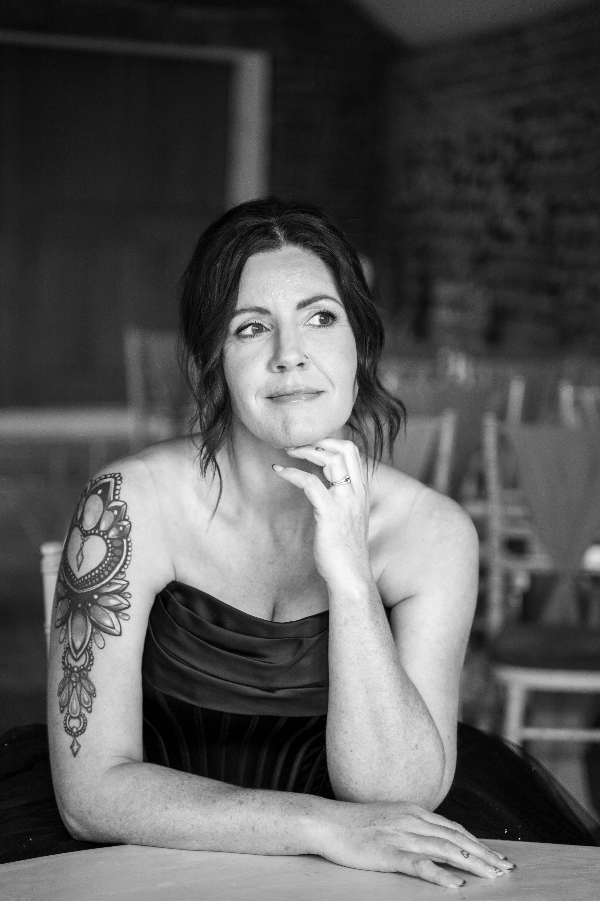 Black and white photograph of a woman with chin resting on hand, shoulder strap dress, and floral tattoo on her arm, sitting at a table with chairs in the background.