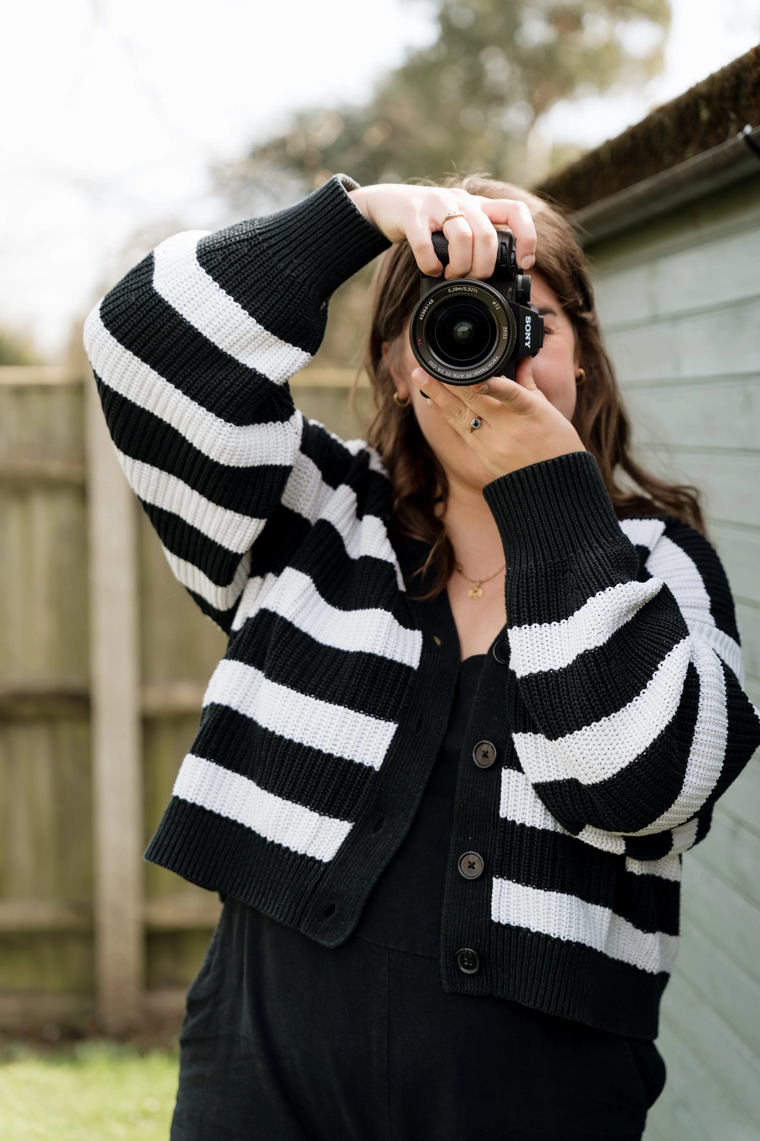 Person wearing a black and white striped cardigan, taking a photo with a Sony camera outdoors near a fence and trees.