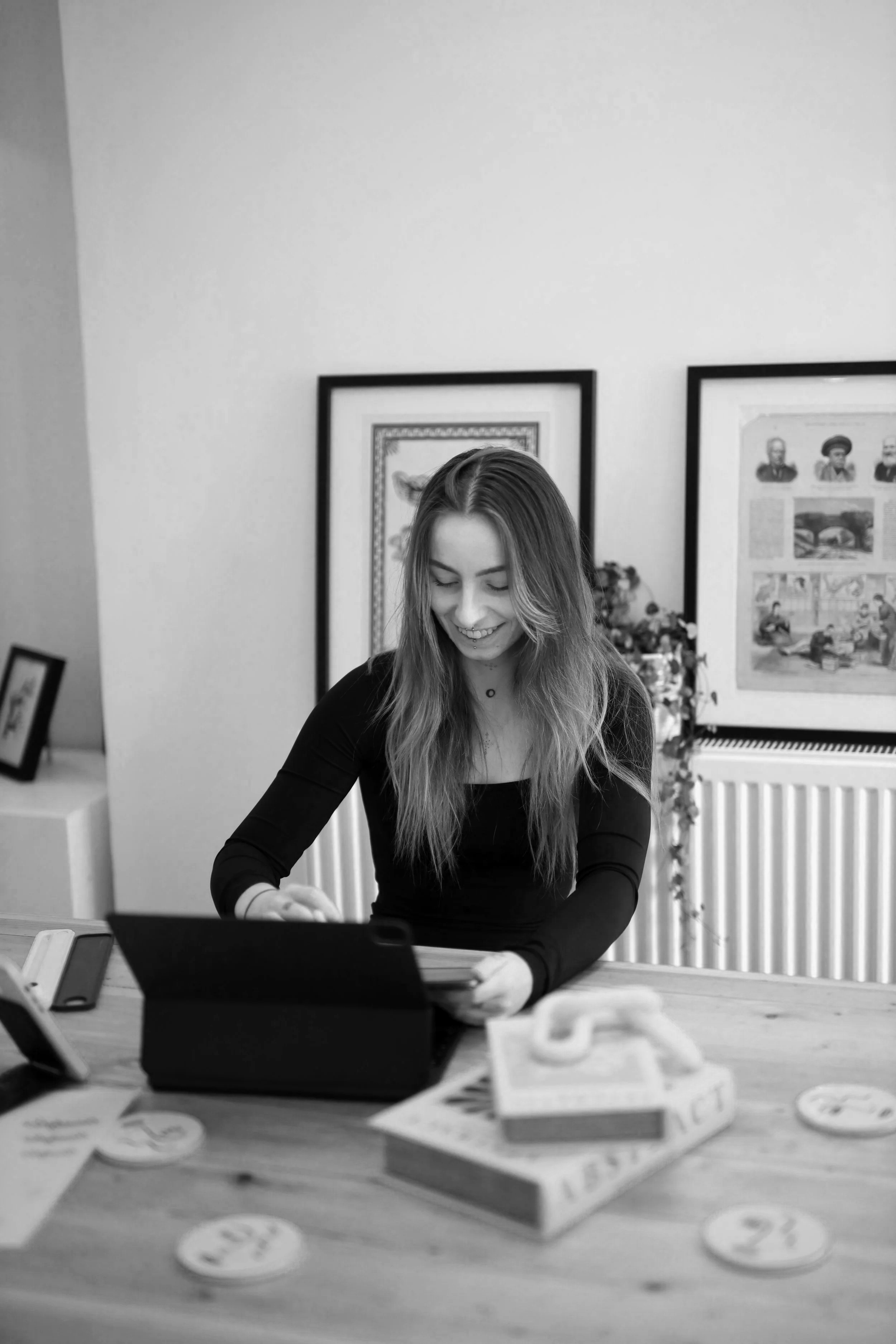 A smiling young woman with long hair, wearing a black top, is sitting at a wooden table looking at a tablet. There are various books and papers on the table, and artwork on the walls behind her.
