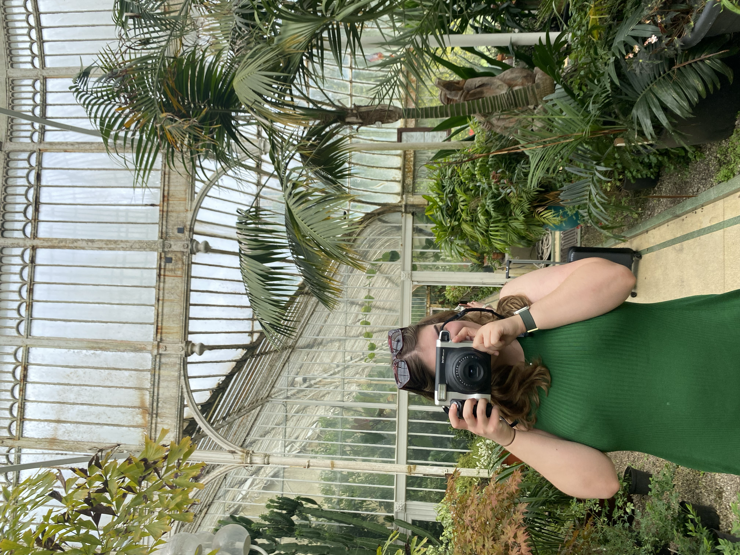 A woman in a green dress taking a photograph with a camera inside a greenhouse filled with various tropical plants and greenery.