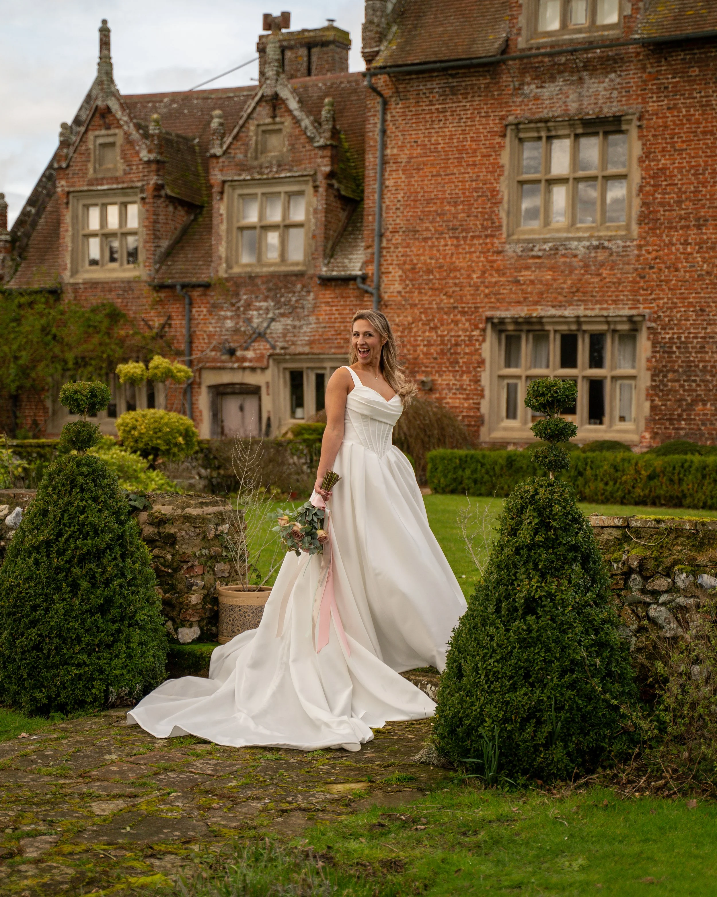 A woman in a wedding dress standing outdoors in front of a brick building with multiple windows and a tariff roof, surrounded by small topiary bushes and greenery.