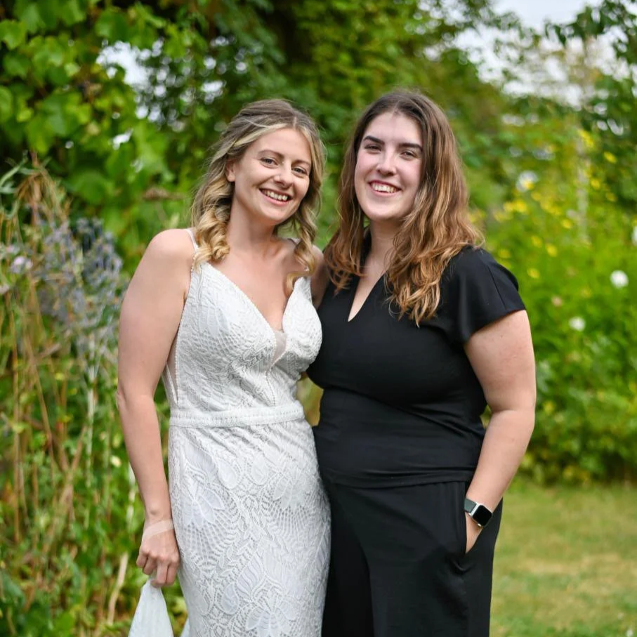 Two women standing outdoors in a garden, smiling at the camera. One is wearing a white lace dress, and the other is in a black dress with short sleeves.