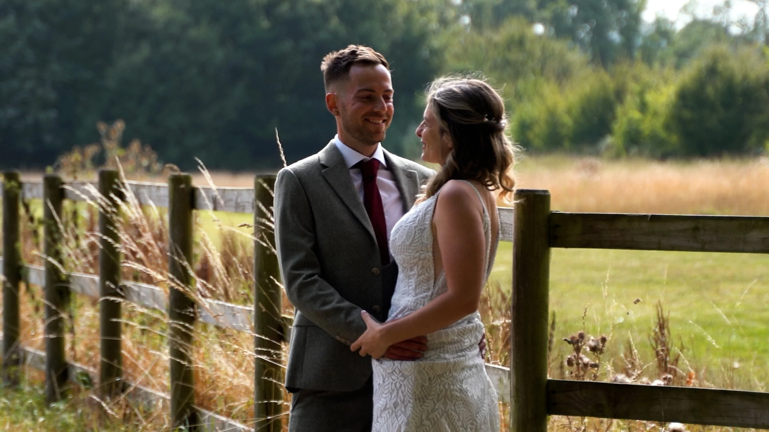 A couple, a man in a suit and tie and a woman in a white dress, standing close together outdoors behind a wooden fence, smiling at each other in a field with trees in the background.