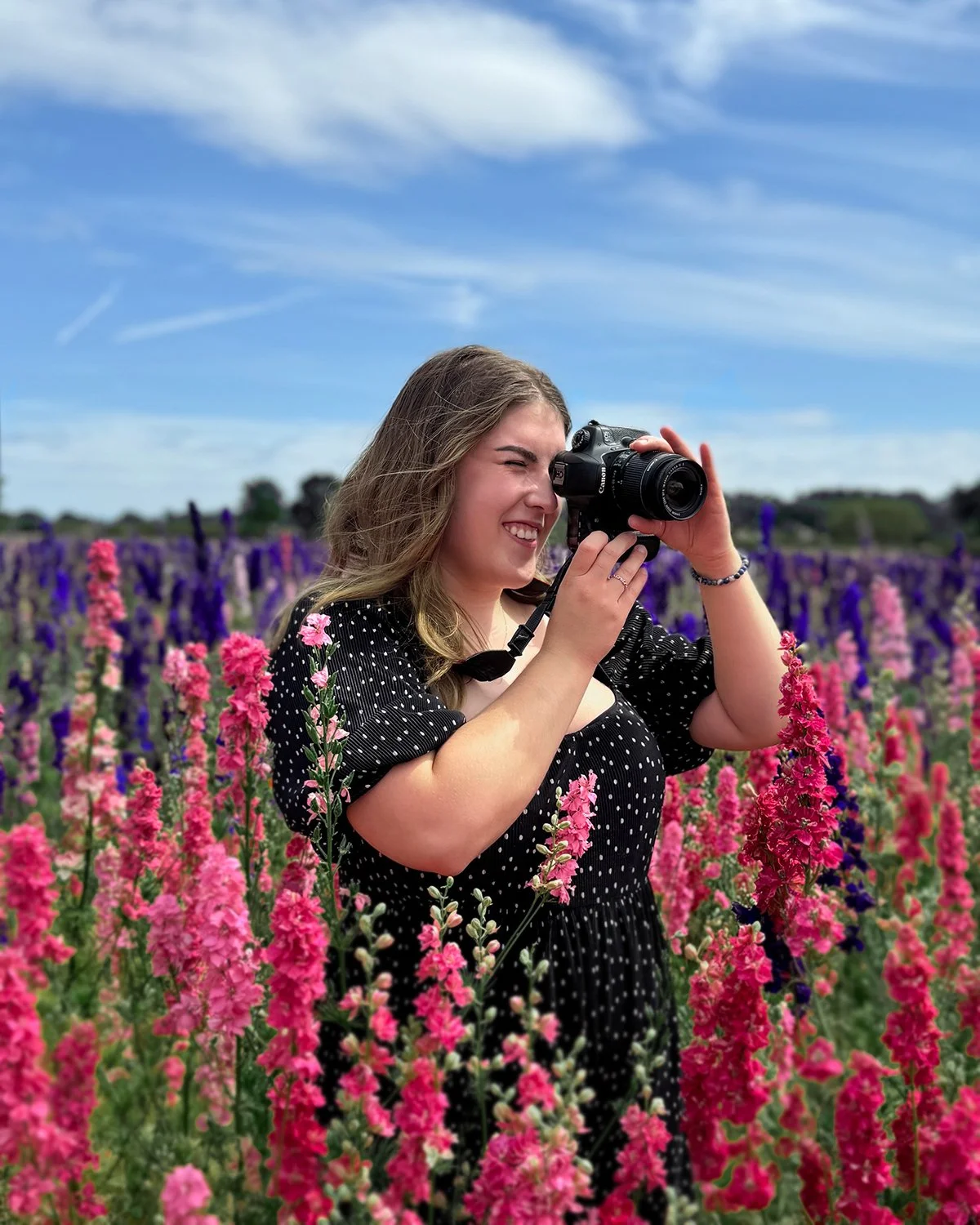A woman with light brown hair wearing a black polka dot dress, smiling and taking a photograph with a camera in a colorful flower field under a partly cloudy sky.