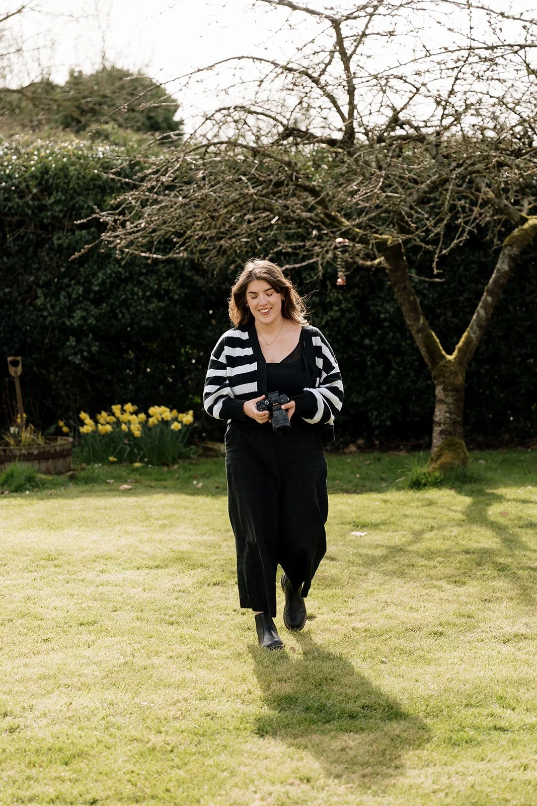 A woman walking outdoors on a grassy lawn, holding a camera, with a tree and bushes in the background.