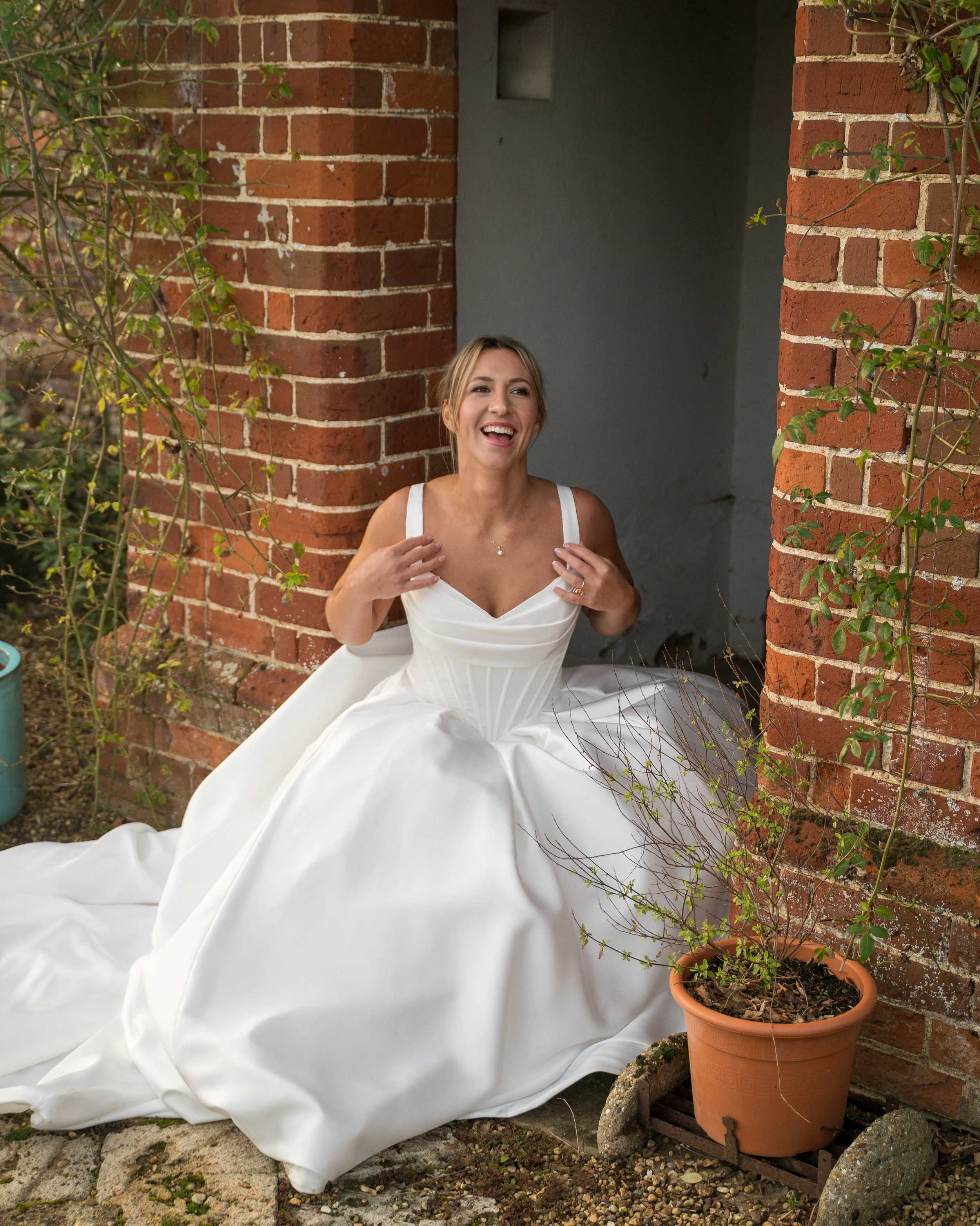 A woman in a white wedding dress smiling and adjusting her dress while sitting outdoors near brick pillars and a potted plant.