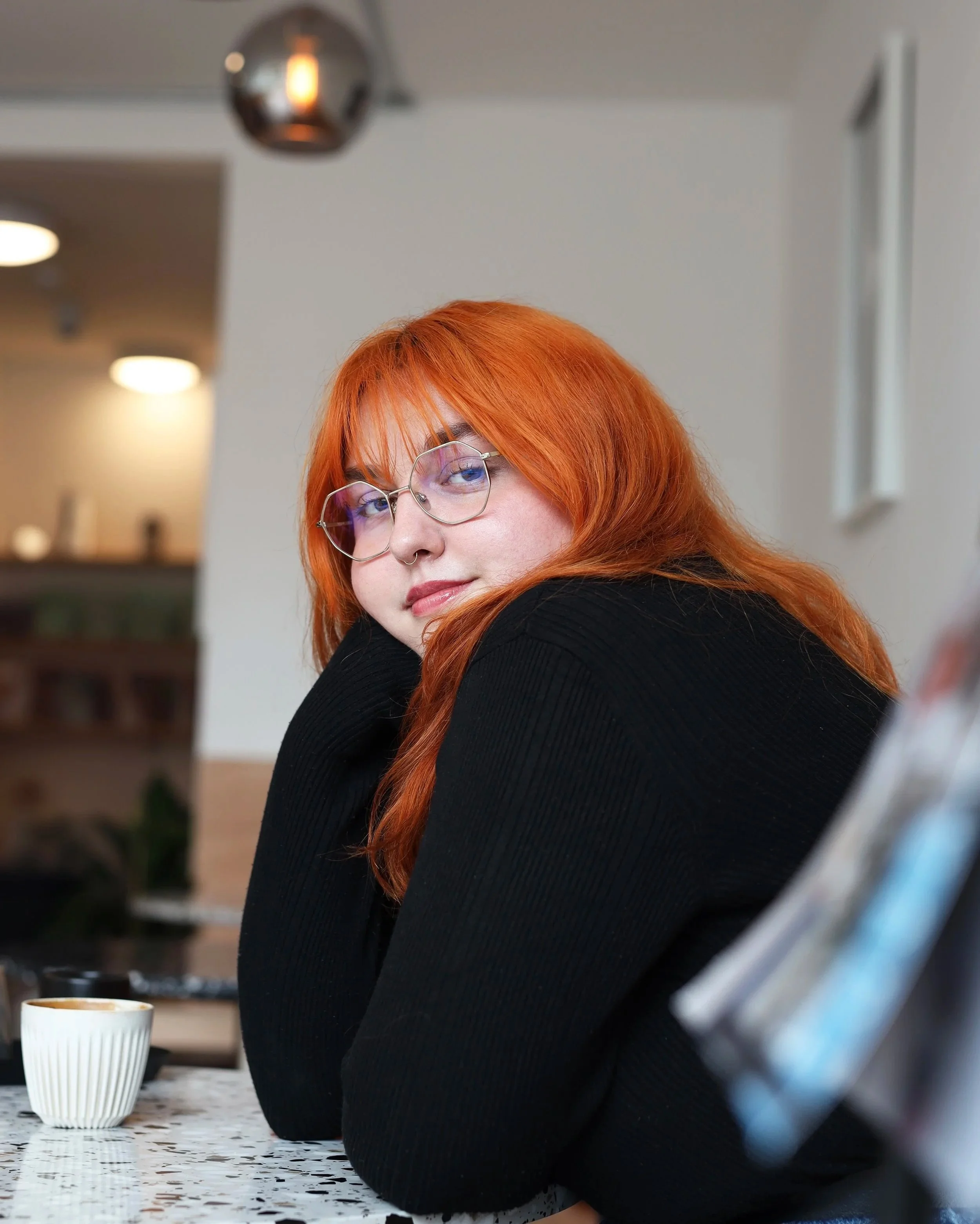 Close-up of a woman with red hair and glasses sitting at a table with cups, in a home or studio kitchen.