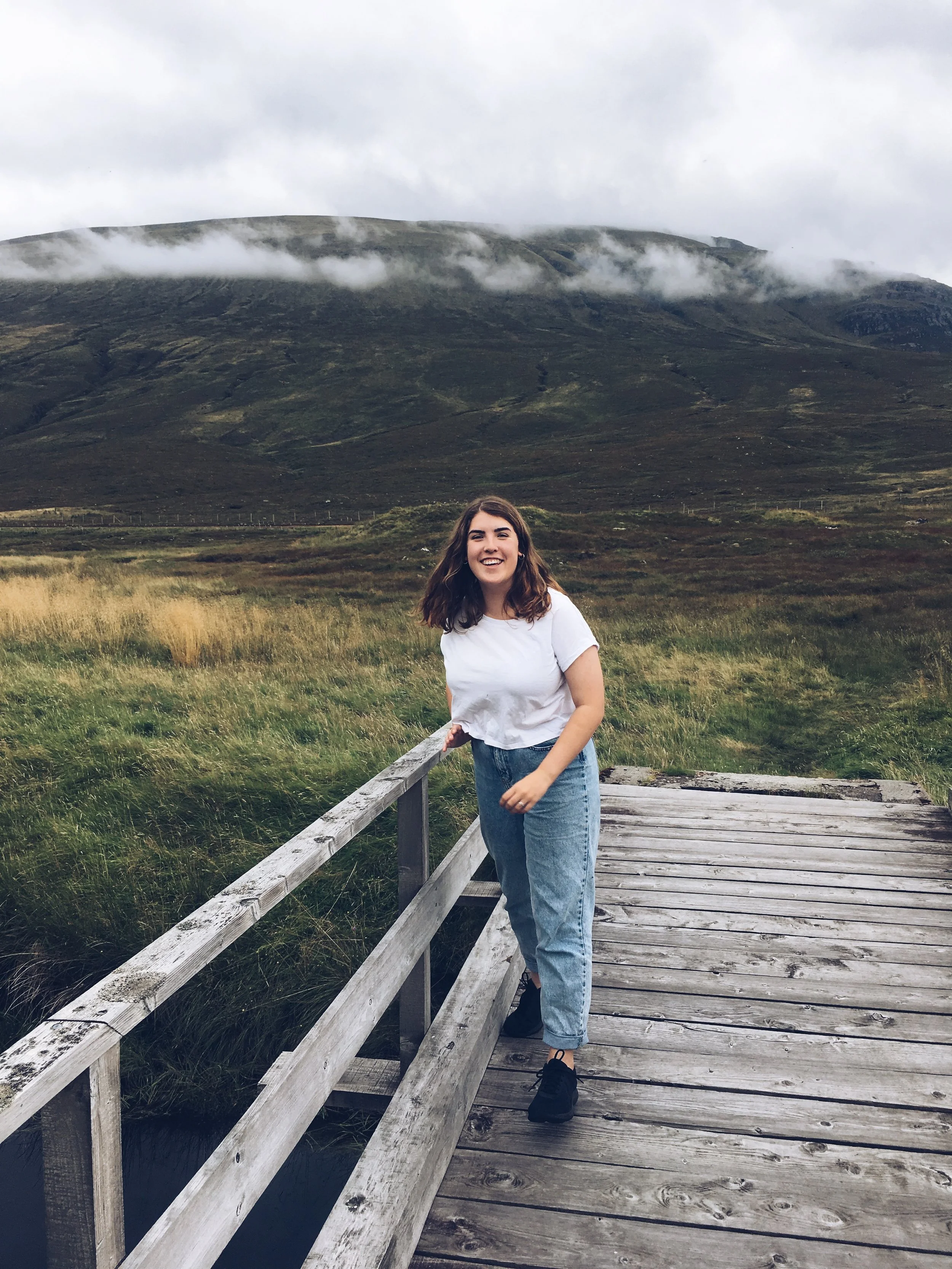 A young woman with brown hair, wearing a white t-shirt, blue jeans, and black shoes, standing on a wooden bridge. She is smiling and holding onto the bridge railing, with a mountain landscape and cloudy sky in the background.